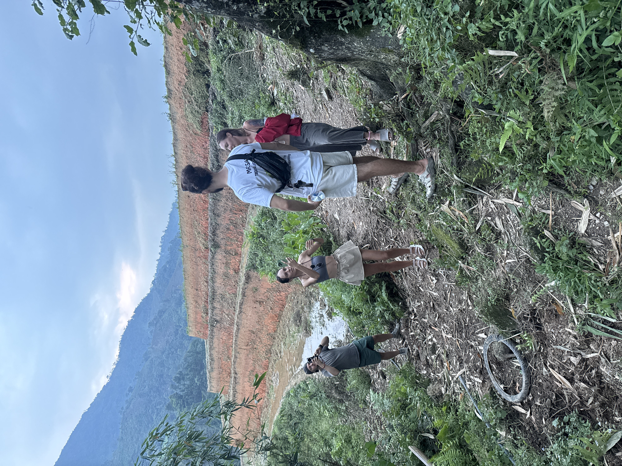 Five individuals are captured on a rural path traversing a mountainous, terraced landscape in Nậm Dịch Commune, Vietnam.
In the foreground, a man with dark hair, wearing a white t-shirt with partially visible text "MKSA TREING ASS", beige shorts, and a black fanny pack, holds a water bottle. He stands with his back mostly to the camera. Behind him, a woman in a red top and dark pants also faces away. Further along the path to the left, a woman in a grey crop top, light skirt, and white sandals wears headphones and looks towards the camera with a raised hand. Beside her, a man in a grey t-shirt, dark shorts, and a dark cap is seen holding a camera to his eye, his reflection visible in a muddy puddle or water-filled section of the path. A fifth person is partially obscured by foliage in the far background on the right.
The surroundings feature lush green vegetation, including ferns and bamboo, lining the path. To the left, distinct terraced fields with reddish-brown soil ascend the hillside. In the distance, rolling mountains covered in dense vegetation are visible under a bright, hazy sky. An abandoned tire lies on the ground to the right of the path. The group appears to be hiking or trekking, taking a break to observe and photograph the scenic environment. The time of day is daytime, with clear but somewhat hazy weather.