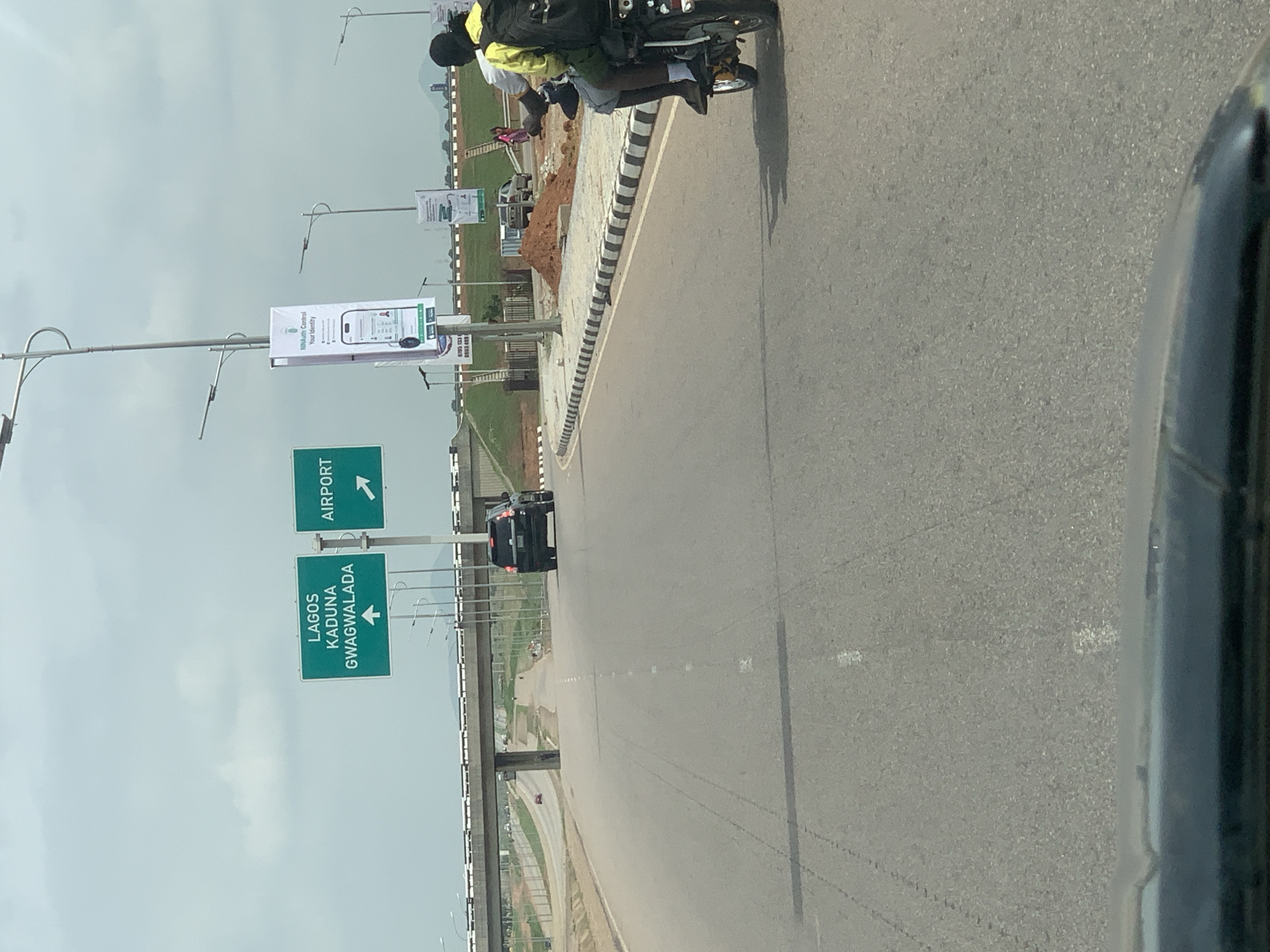 This daytime photograph, likely taken from inside a vehicle, captures a major road scene in the Immigration Quarters area of Nigeria under an overcast sky. The road stretches into the distance, with a clear asphalt surface marked by faded white dashed lines. On the right side of the road, a motorcycle, commonly known as an "okada," carries a driver and a passenger, both wearing casual attire, and appears to be moving away from the camera. A striped concrete barrier separates the main carriageway from a shoulder or median where construction activity, including a pile of reddish dirt, is visible.
On the left side of the road, several tall street light poles are visible, from which banners and road signs are suspended. Two prominent green road signs provide directions: one indicates "LAGOS KADUNA GWAGWALADA" with a left arrow, and another, positioned above it, points right to "AIRPORT." Further along the left side, a white banner prominently displays the "NIMcApp" logo, along with the slogan "Control Your Identity" and an illustration of a smartphone. Another smaller banner below it shows partially visible phone numbers, including "0700 331" and "0800 68." In the mid-distance, a black car is driving on the road, and an overpass or bridge structure spans across the scene, with more traffic visible beneath it. The overall impression is one of a busy, developing urban highway.