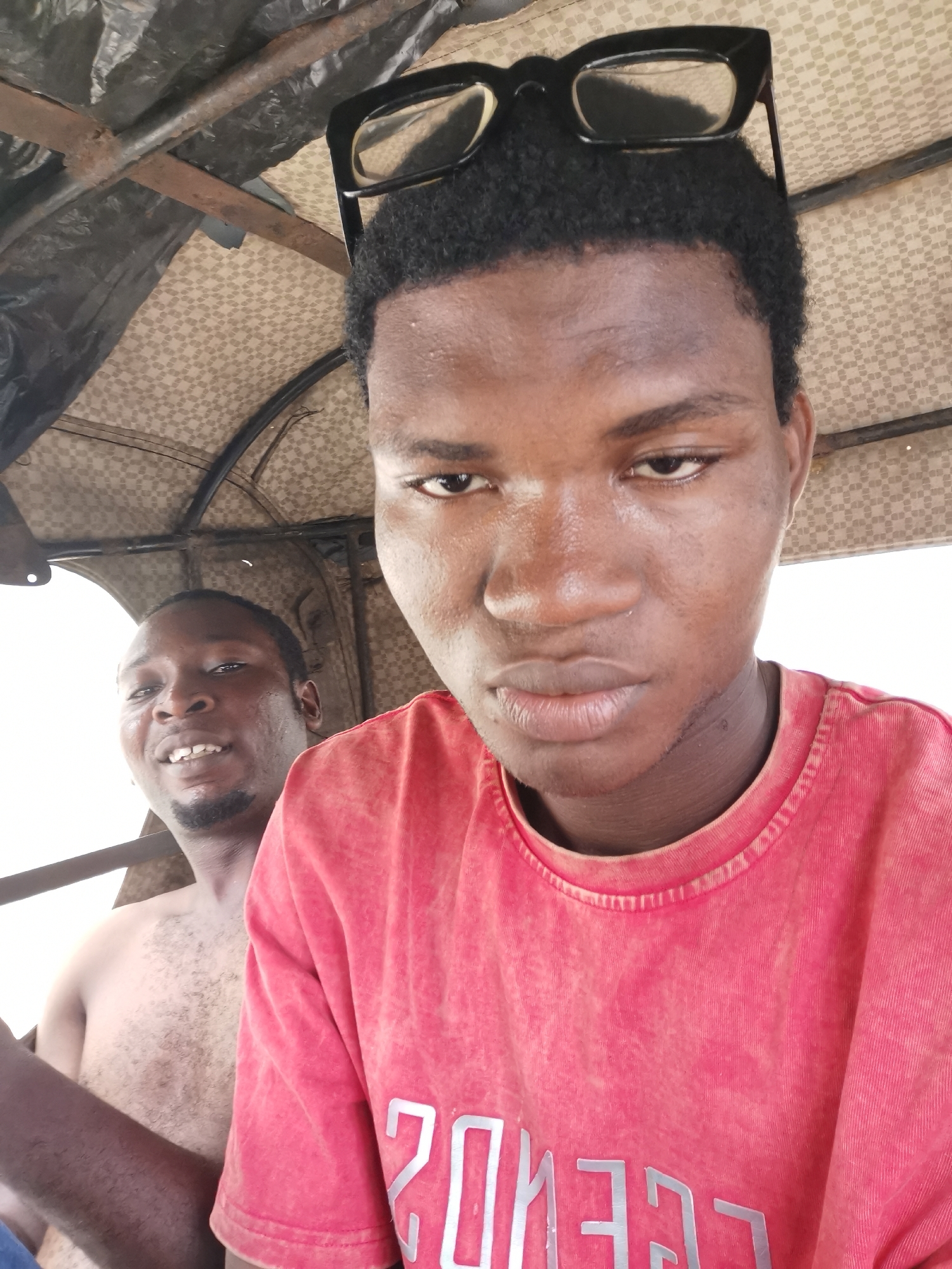 Two men are seated inside a vehicle, possibly a motorized tricycle taxi known as a "Keke Napep" in Lagos, Nigeria. The man in the foreground wears a red t-shirt and has sunglasses perched on his head. He is looking directly at the camera with a serious expression. In the background, another man is visible, bare-chested, smiling slightly. The interior of the vehicle has a patterned roof and metal framework. The lighting appears to be natural daylight. The location is likely Lagos, Nigeria, based on the presence of the Keke Napep, a common mode of transportation in the city.