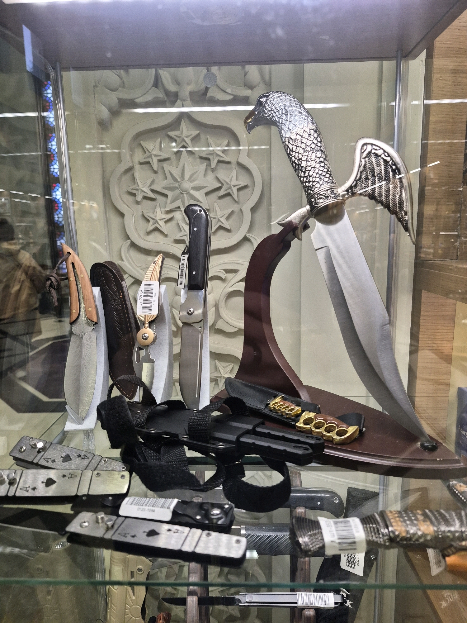 A display case in a shop contains various knives and related items. The case has glass shelves and is illuminated from above. In the background, a decorative panel with a star motif is visible.
The prominent item is a large decorative knife with a blade and a handle shaped like an eagle's head and wings. This knife is displayed on a dark brown stand. To the left of the large knife, three smaller knives are displayed upright on stands. The leftmost of these has a wooden handle and a Damascus steel blade. The middle knife appears to be a folding knife with a brass-colored object attached to its handle, possibly a keychain or decorative element. The rightmost of these three knives is a fixed-blade knife with a black handle.
Below these, on the glass shelf, is a black sheath containing brass knuckles. Several other knives are visible on the lower shelves, including folding knives with metal handles decorated with spade suit symbols. Another knife with a textured handle and a barcode label is also present. Some of these knives are secured with black straps.
The context suggests this is a retail environment, likely a weapons or collectibles shop. The presence of decorative and functional knives, along with brass knuckles, points towards items related to combat, self-defense, or collecting. The background hints at an indoor setting with some decorative elements, possibly a specialized store. No people are clearly visible, though reflections suggest the presence of a photographer or onlookers.
The image does not provide any specific geographical markers to determine its location within Moscow, Russia.