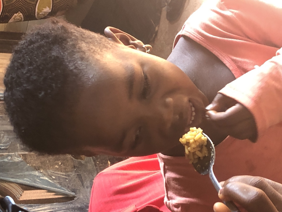 A young girl with dark, curly hair eats from a spoon. She is angled to the left of the frame and wears a pink long-sleeved shirt. Her eyes are partially closed, and she smiles, showing her teeth as she brings food to her mouth. The spoon holds rice or grains of a yellow-brown color. A hand partially visible in the foreground appears to be holding the spoon. The background includes blurred items, possibly furniture and clothing, in shades of black, brown, and beige.