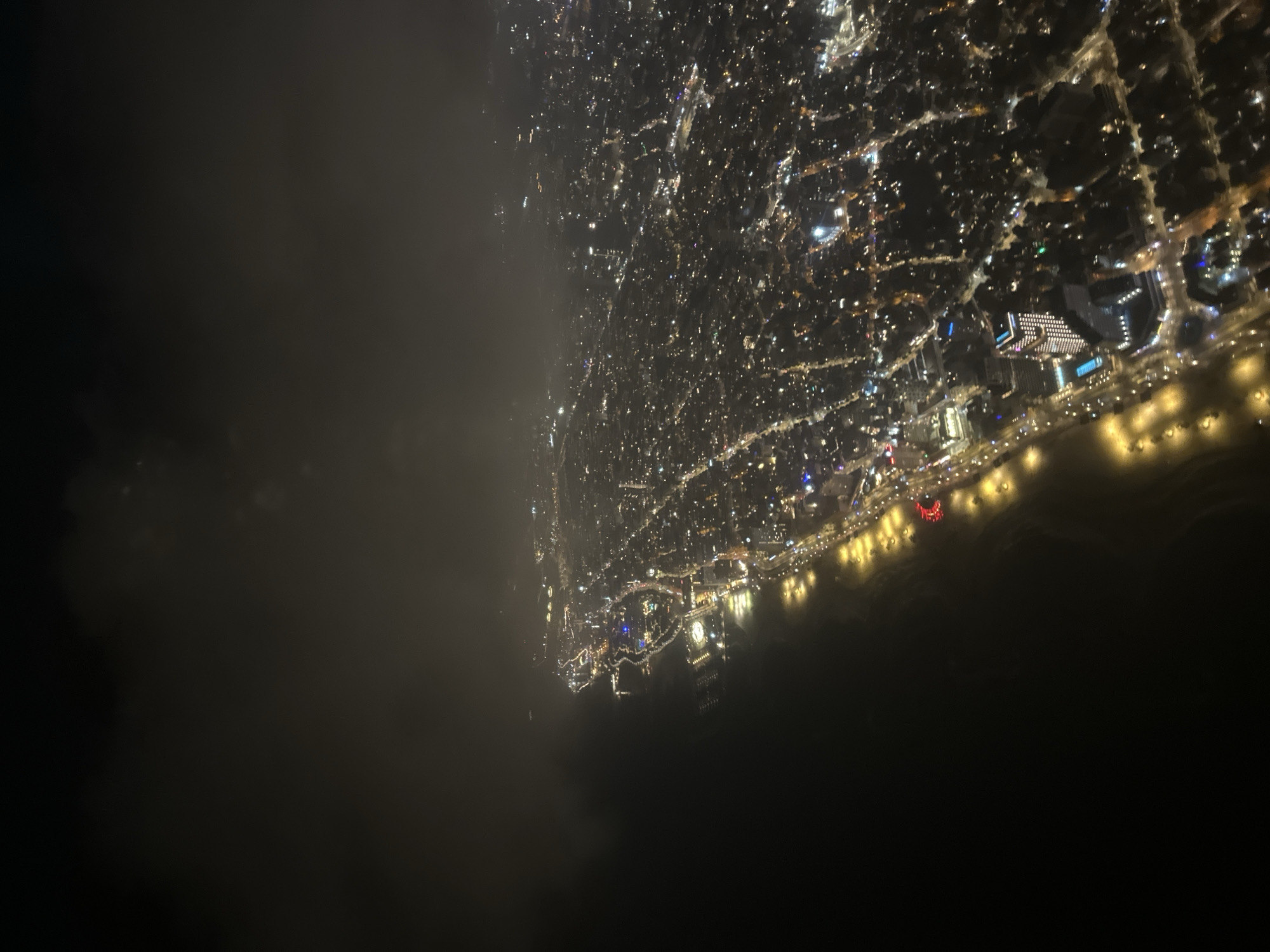 The image is an aerial nighttime view of Tel Aviv, Israel. The city lights illuminate the coastline. Several prominent buildings and landmarks are visible along the shore, including hotels, entertainment areas, and possibly the port. The sea appears dark and still. Clouds or fog obscure the left side of the image, creating a contrast between the brightly lit city and the darkness of the surrounding environment. No people or interactions are discernible at this scale.