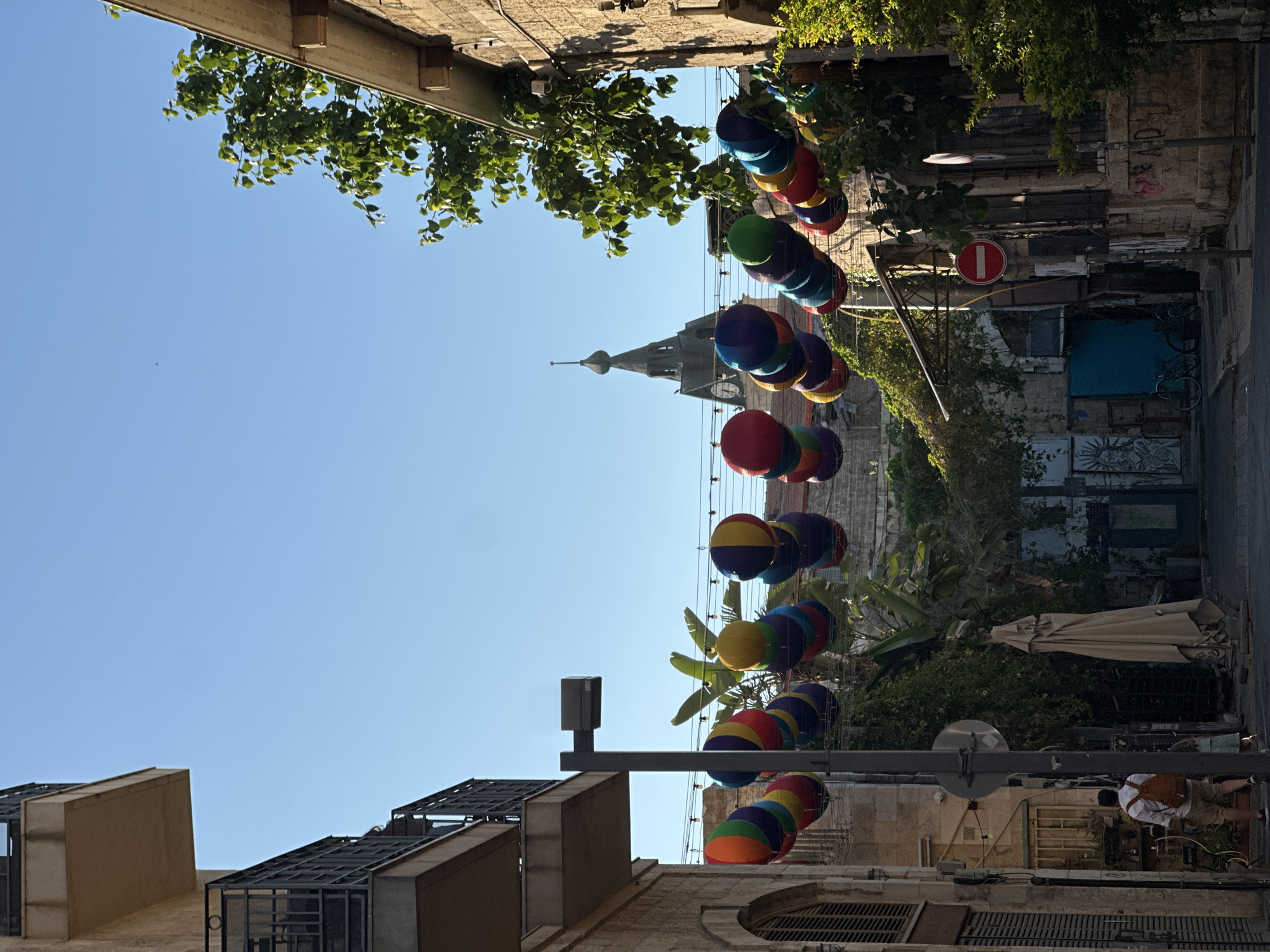 The image depicts a street scene in Tel Aviv, Israel, likely in the Neve Tzedek neighborhood, known for its narrow streets and historic architecture. The street is lined with buildings, some with balconies and stone facades. Overhead, a string of colorful, balloon-like decorations hangs across the street, adding a festive atmosphere. Sunlight casts shadows, indicating it is likely daytime.
In the foreground, on the left side of the street, a man with a backpack walks away from the camera, accompanied by a child. They are walking on a paved sidewalk. On the right side of the street, lush green trees and foliage spill over the walls of the buildings. A red and white circular "no entry" sign is visible on a pole. Further down the street, a church spire with a clock is visible against the clear blue sky. The street appears to be pedestrian-friendly, with bicycles parked in some areas. The overall impression is one of a charming and vibrant urban environment.