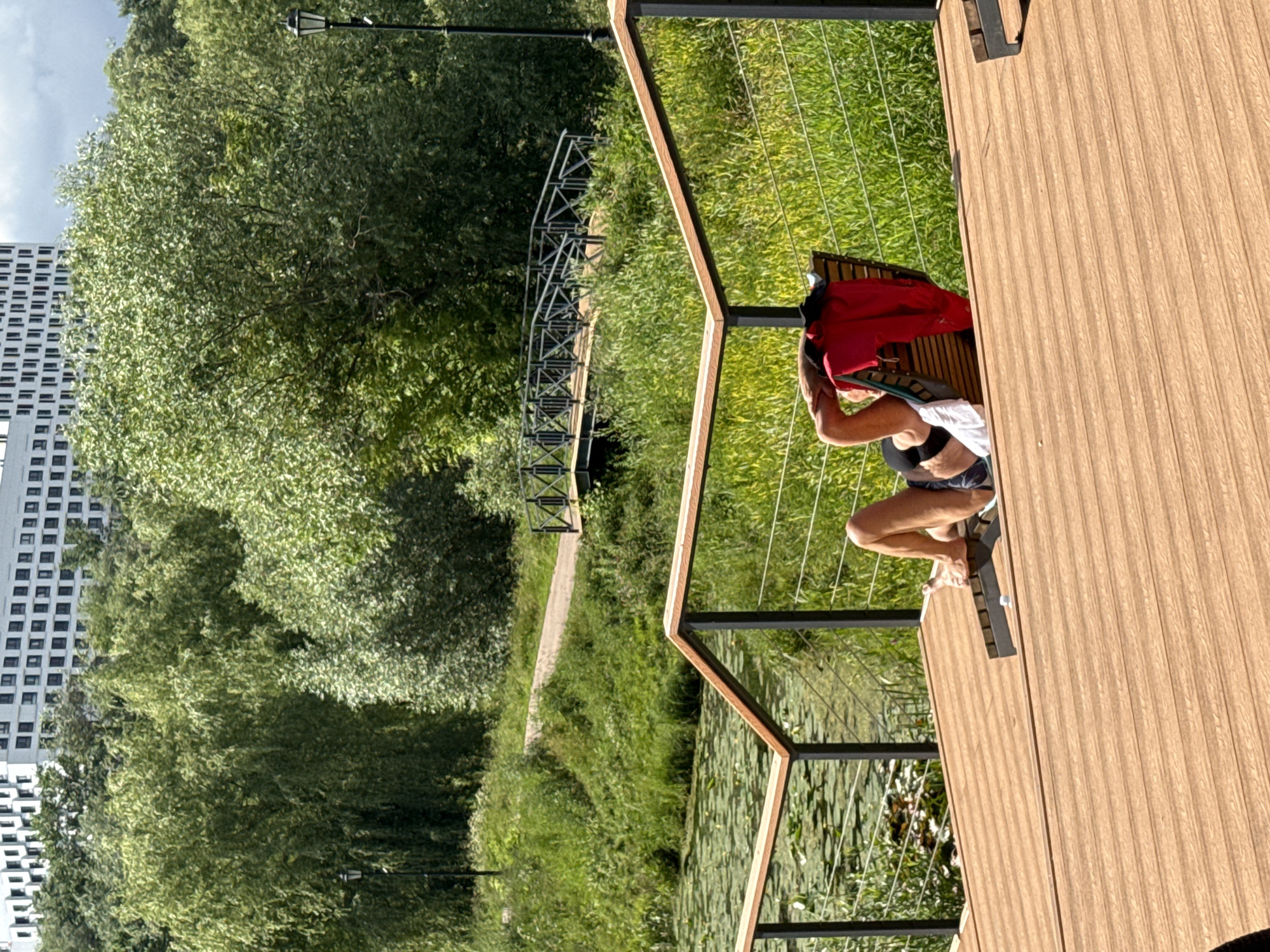 The image shows a person sitting on a wooden deck overlooking a body of water with lily pads. The person is wearing a red shirt and patterned shorts. To the left of the deck, there is a grassy area with trees and a walking path leading to a metal bridge. In the background, a tall, modern white building with many windows is visible, suggesting an urban or suburban setting. The scene appears to be set during the daytime, with bright sunlight casting shadows, indicating fair weather. The overall atmosphere is relaxed, with the person seemingly enjoying the outdoor view. The presence of the modern building and lush greenery suggests this might be a park or recreational area within or on the outskirts of Moscow, Russia. There is no visible text in the image.