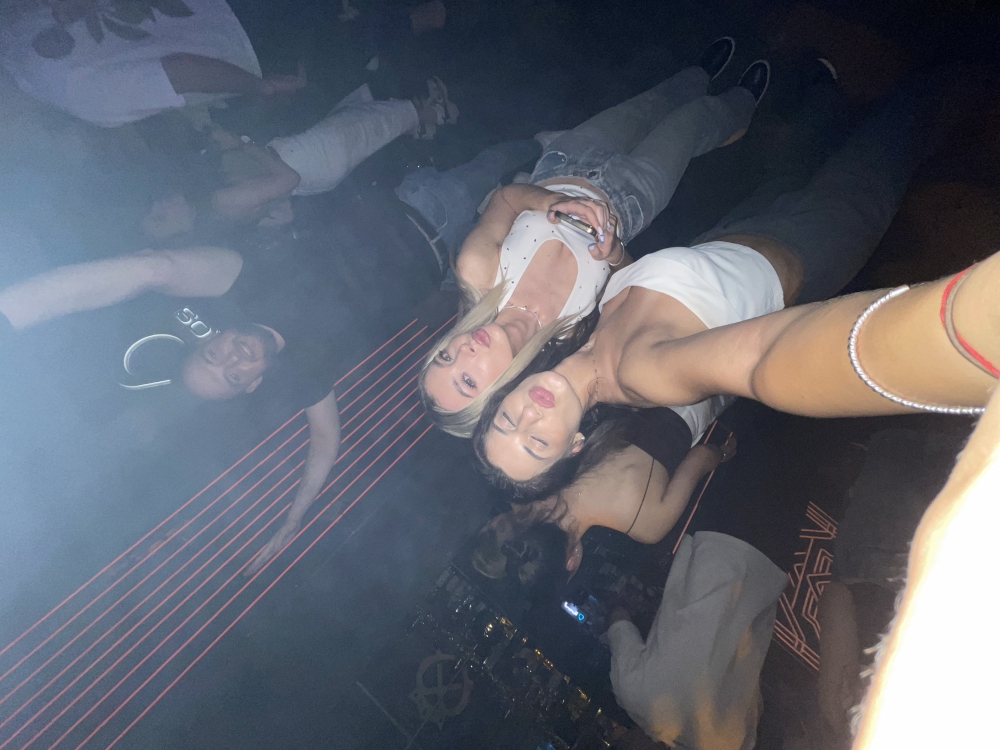 The image depicts an indoor, dimly lit, social gathering, likely a nightclub or bar, in the city of Unknown, Unknown. The scene is captured from a low-angle selfie perspective, looking upwards and diagonally.
In the immediate foreground, the right arm and shoulder of an individual with tan skin are visible, wearing a white one-shoulder crop top. A silver beaded bracelet and a red string bracelet adorn the wrist.
Centered in the mid-ground are two women posing for the camera. The woman on the right has long blonde hair, is wearing a white halter-neck crop top with embellishments, light blue ripped jeans, and dark shoes. She holds a gold-colored smartphone with both hands and looks directly at the camera with slightly parted lips. The woman on the left has long dark hair, is wearing a black strappy backless top, and dark pants. She is making a "kissing" face with closed eyes towards the camera.
In the background, multiple individuals are present. Towards the upper left, a man with a beard and dark hair, wearing a black t-shirt, smiles at the camera with his left arm raised. Behind him, a partial illuminated sign with the letters "D" and "SO" is visible. To the upper right, the torso and legs of an individual wearing a white t-shirt with a dark leaf pattern are visible. Another person in the middle-right background wears a light-colored dress or skirt. Further left, what appears to be a bar area with bottles and glassware is discernible. On the floor or a low surface in the lower-middle, a geometric illuminated pattern resembling letters such as "FAVE" is present.
The environment is characterized by atmospheric haze or smoke, and red laser lines diagonally traverse the upper and mid-background. The overall lighting is low, contributing to a nighttime indoor ambiance.