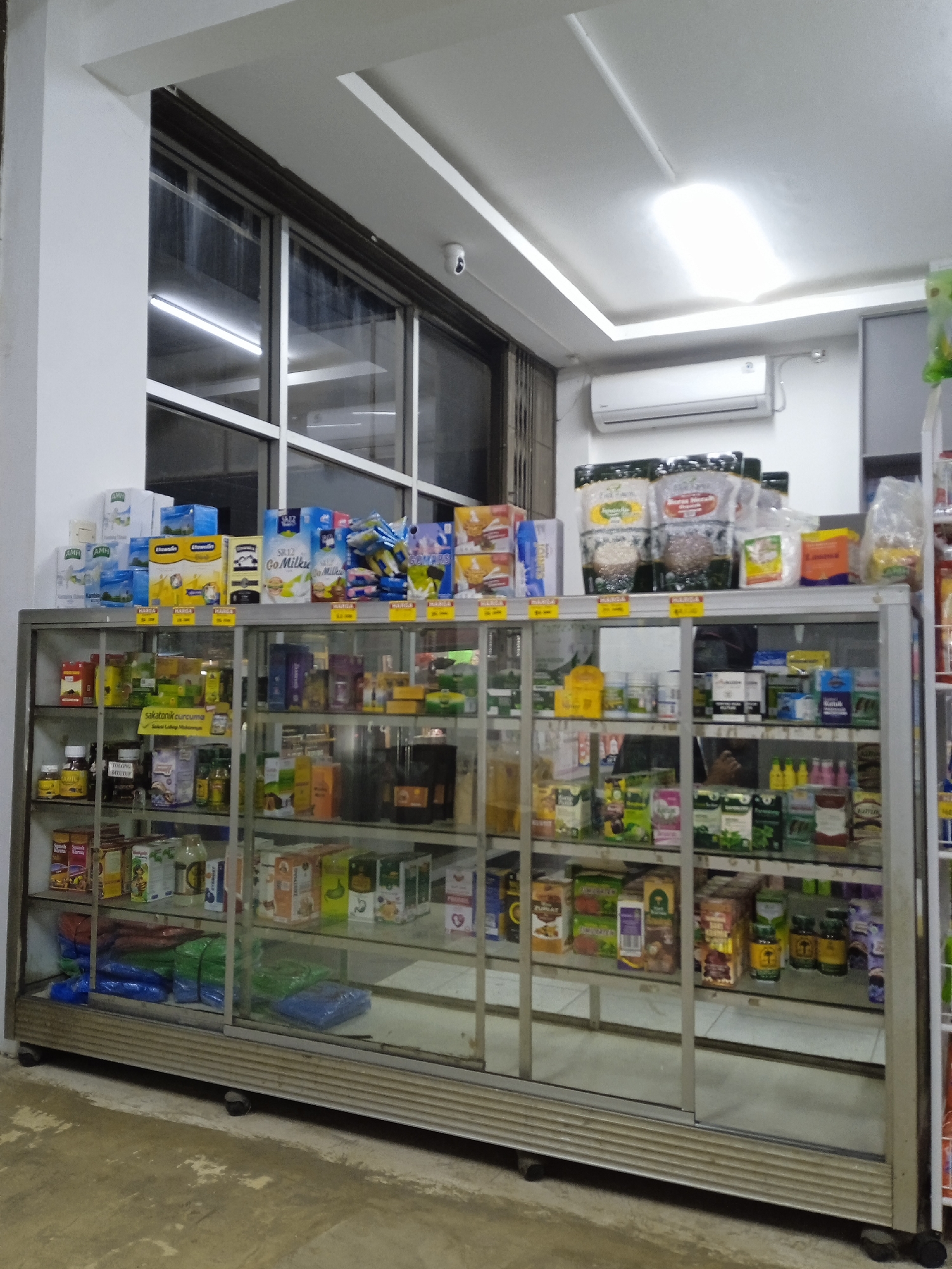 This image captures the interior of a small shop or mini-mart, likely in Kebonsari, Indonesia. The central focus is a large, glass display cabinet filled with various packaged goods.

On top of the cabinet, and inside its shelves, are numerous products. These include boxes of milk powder or supplements (e.g., "AMH", "SR12 Go Milku", "Elhassan"), bags of what appear to be food ingredients or health supplements ("Pure Honey", "Susu Kambing Etawa"), and other packaged items. Many products feature yellow price tags marked "HARGA" (Indonesian for "price"). Brand names like "sakatonic curcuma" are also visible, indicating health or herbal products. The lower shelves of the cabinet contain more boxed items, bottles, and neatly folded plastic bags in blue and green.

In the background, a large window with white frames allows natural light into the space, though overhead ceiling lights, including a large rectangular panel light, are also illuminated. An air conditioner unit is mounted on the wall below the ceiling, to the right of the window. A small security camera (CCTV) is visible near the top right corner of the window frame. The floor is made of a light-colored, textured concrete or tile.

No people are visible in the scene, and there is no discernible activity other than the static display of merchandise. The lighting suggests it could be daytime, with artificial lighting supplementing the natural light. The overall impression is that of a tidy, functional retail space.