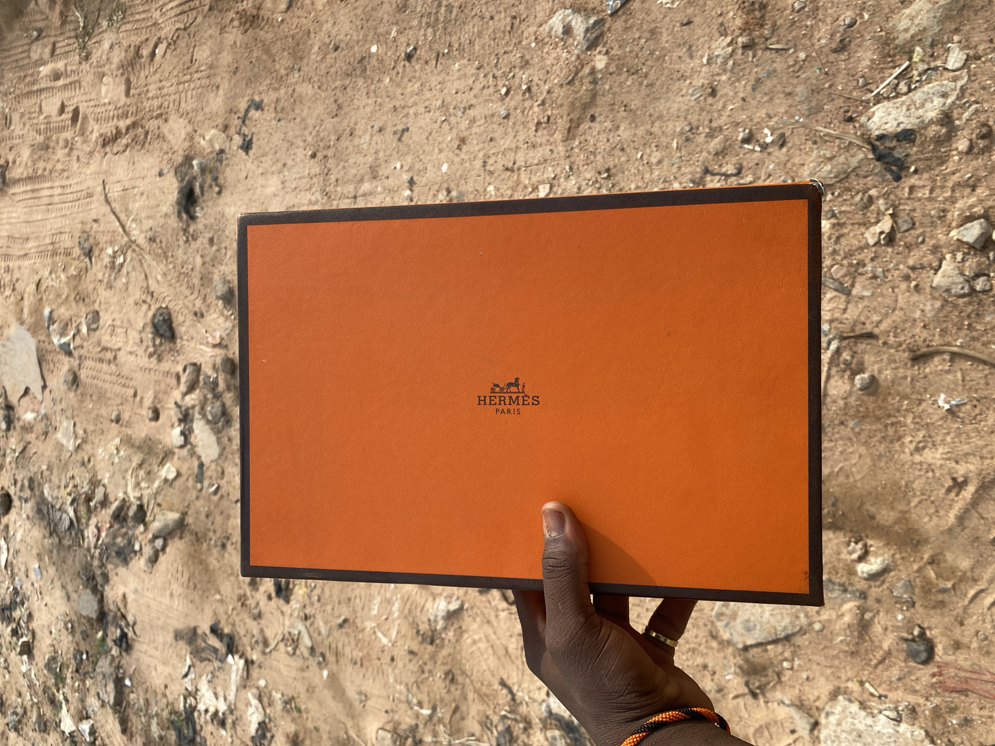 An overhead shot shows a person's left hand holding an orange Hermès box against a backdrop of dusty, uneven ground in Jalingo, Nigeria. The box is rectangular and appears to be made of stiff cardstock. The Hermès logo, featuring the brand name and "PARIS" in black text alongside an equestrian crest, is visible on the box's surface. The hand is dark-skinned and wears a beaded bracelet on the wrist and a ring on one finger. The ground is composed of loose sand, small stones, and scattered debris, with tire tracks imprinted on its surface.