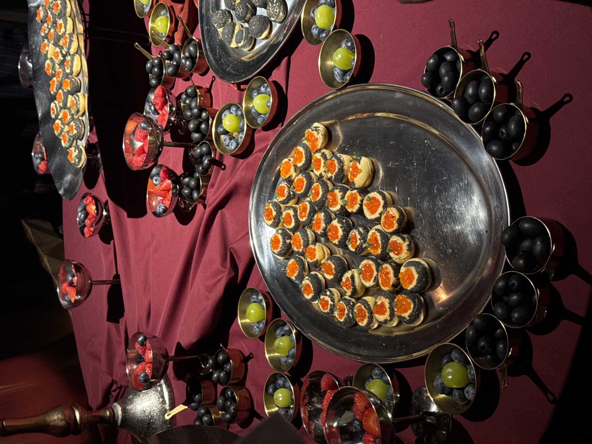 The scene depicts an indoor catering display set on a table draped with a deep reddish-maroon tablecloth, located in the city of Yerevan, Armenia. The lighting is dim, casting reflections on metallic surfaces.
In the foreground and center, a large, oval, reflective silver platter presents approximately 35-40 small, round canapés. These canapés feature light-colored pastry bases, some with a dark outer coating, each topped with a dollop of light-colored cream and bright orange fish roe. To the right and slightly in the background, a smaller, elevated, dark-colored oval platter holds similar small pastry items, some black and textured, others light-colored, but without the roe topping.
Numerous small, brass-colored mini-pots with handles are distributed across the table. Many of these pots are filled with whole black olives. Others contain blueberries alongside a single green grape. Several reflective, metallic goblet-style bowls are also present, filled with mixed fresh berries, specifically red strawberries, red raspberries, and dark blue blueberries, placed at various points on the table.
On the far left, part of a tall, ornate metallic stand, resembling a candelabra base, is visible. No individuals are clearly depicted, though a partial dark sleeve or garment is visible at the far left edge of the frame. The scene is static, showcasing a prepared food arrangement.