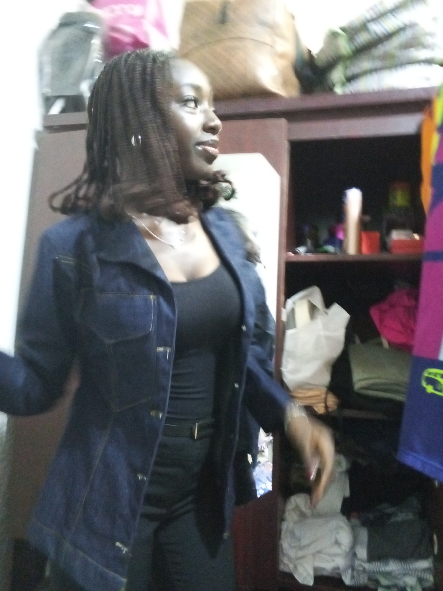 A woman stands indoors, likely in a private residence. She is wearing a dark tank top, a navy blue denim jacket, and dark pants. Her hair is styled in braids and she has hoop earrings. She is looking to her right. Behind her is a dark-colored wooden wardrobe or shelving unit filled with various items, including folded clothing, bags, and toiletries. There is additional storage on top of the unit. There is no definitive evidence to establish this is the city of Abuja, Nigeria.