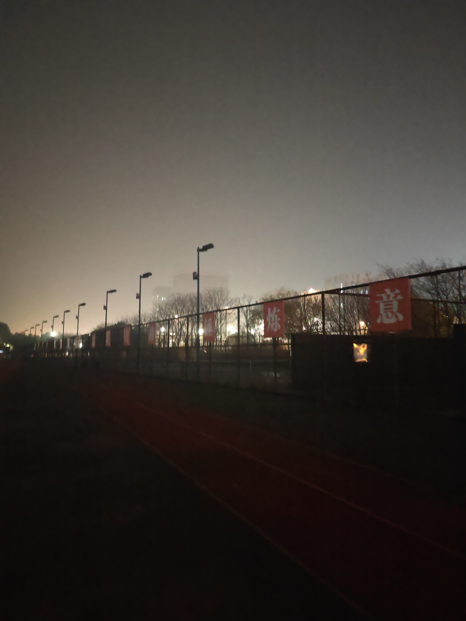The image shows a running track at night in Jiangning District, China. The track is dark, with red lines indicating lanes. A fence runs alongside the track, and several light posts illuminate the scene. Banners with white Chinese characters on red backgrounds are attached to the fence. Trees and buildings are visible in the background, obscured by the darkness and mist. The sky is overcast. There are no people visible in the image.