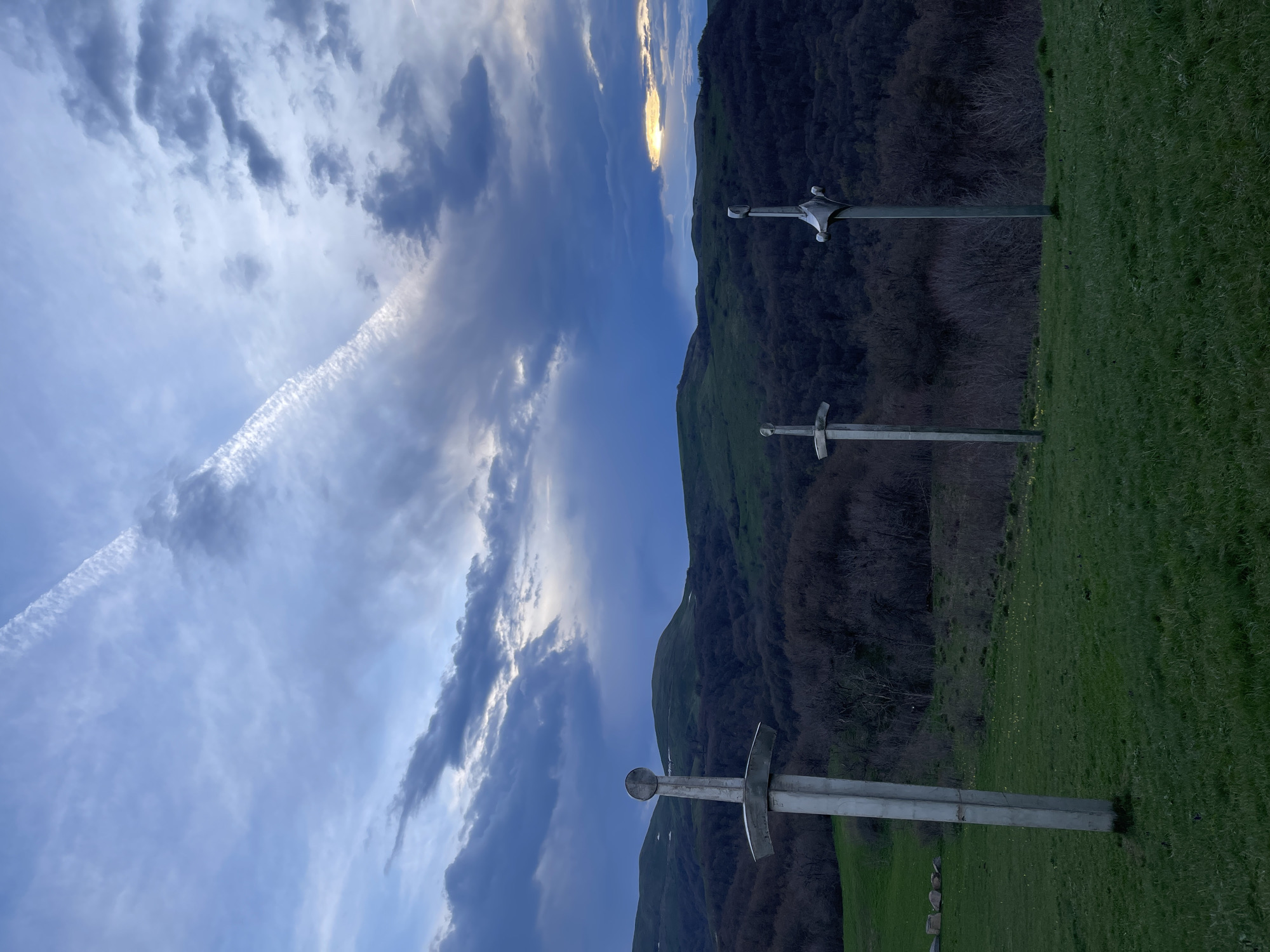 The image shows three large metal sword monuments erected in a grassy field in Shamta, Georgia. Behind the swords is a densely wooded mountain range under a cloudy sky. A contrail is visible in the upper left of the sky. The time appears to be near dusk or dawn due to the lighting and cloud cover.