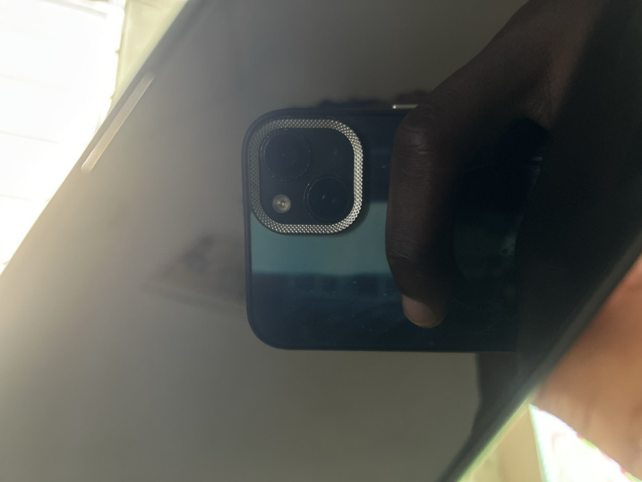 The image displays a reflection of a dark-skinned hand holding a smartphone, visible on a glossy, dark surface. The smartphone is dark green or teal, equipped with a dual-lens camera system and a flash, encased in a silver, textured protective ring. The hand's thumb is visible on the lower right side of the reflected phone. The upper right portion of the reflection shows a bright, diffused light source, possibly a window or overhead light. Faint, indistinct architectural elements and ambient lighting are discernible in the background of the reflection, suggesting an indoor setting. This scene is located in Egbe, Nigeria.