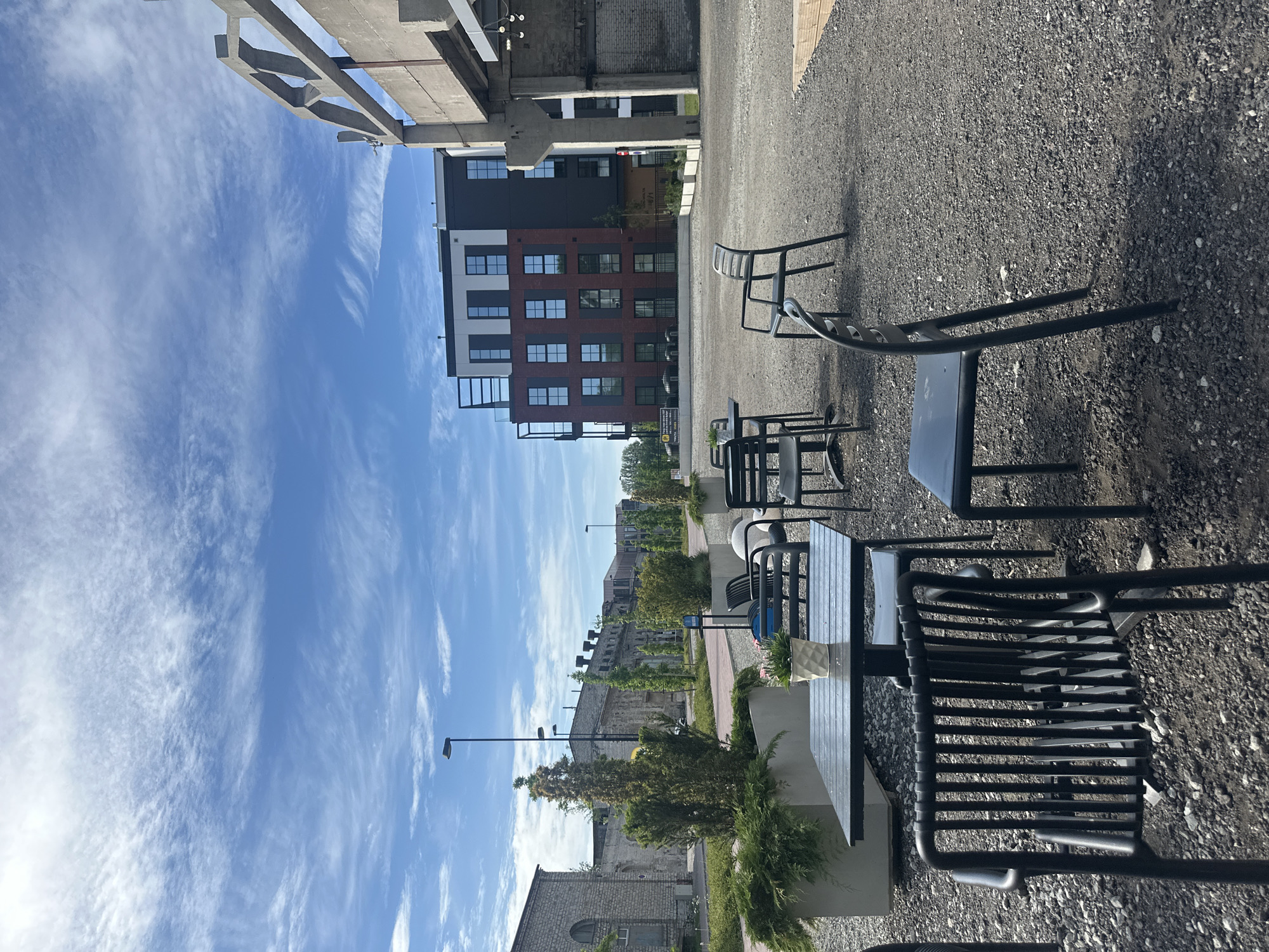 This image depicts an outdoor seating area with tables and chairs set on a gravel surface in the city of Tallinn, Estonia. The scene is situated in front of a modern, multi-story brick building with numerous windows. To the right, a concrete structure, likely part of an elevated roadway or industrial building, casts a shadow. In the background, older, weathered stone buildings line a street with trees and streetlights. The sky above is blue with scattered, wispy clouds. The seating area appears to be unoccupied, with several tables set with chairs and one table featuring a potted plant. The overall impression is of a contemporary urban setting with a blend of modern and older architectural styles.