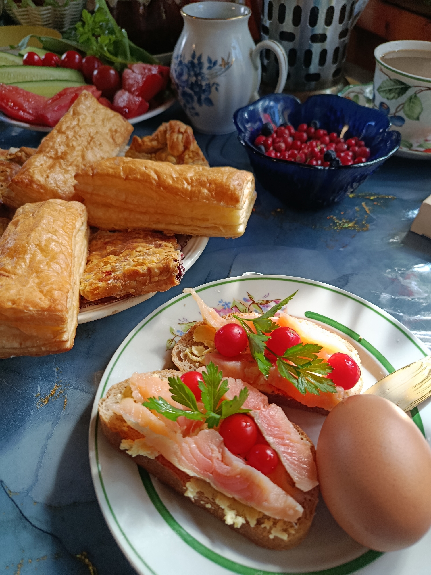 A close-up shot captures a breakfast spread on a blue, marbled surface, likely in a home setting in Verenya, Russia. The scene is bathed in natural light, creating dappled shadows and highlighting textures.
In the foreground, a white plate with a green trim displays two open-faced sandwiches. Each sandwich consists of a slice of dark bread topped with a layer of light-colored spread, thinly sliced smoked salmon, and two small, bright red cherry tomatoes. A sprig of parsley garnishes each sandwich. To the right of the plate, a single, brown, uncooked egg rests. A golden-colored utensil, possibly a fork, is partially visible at the edge of the plate.
Behind the plate, a white dish holds sliced cucumbers, tomatoes, and what appear to be radishes. To the right of this dish, a dark blue, petal-shaped bowl is filled with fresh red currants and a few dark blueberries.
In the background, various baked goods are arranged. Several golden-brown pastries, some appearing flaky and layered, are visible. One rectangular pastry with visible layers is on a small plate. Further back, a white ceramic pitcher with a blue floral design sits next to a metallic object with a grid pattern. A teacup with a green and floral design is also present, filled with a light-brown beverage. The overall impression is a rustic and appetizing breakfast.