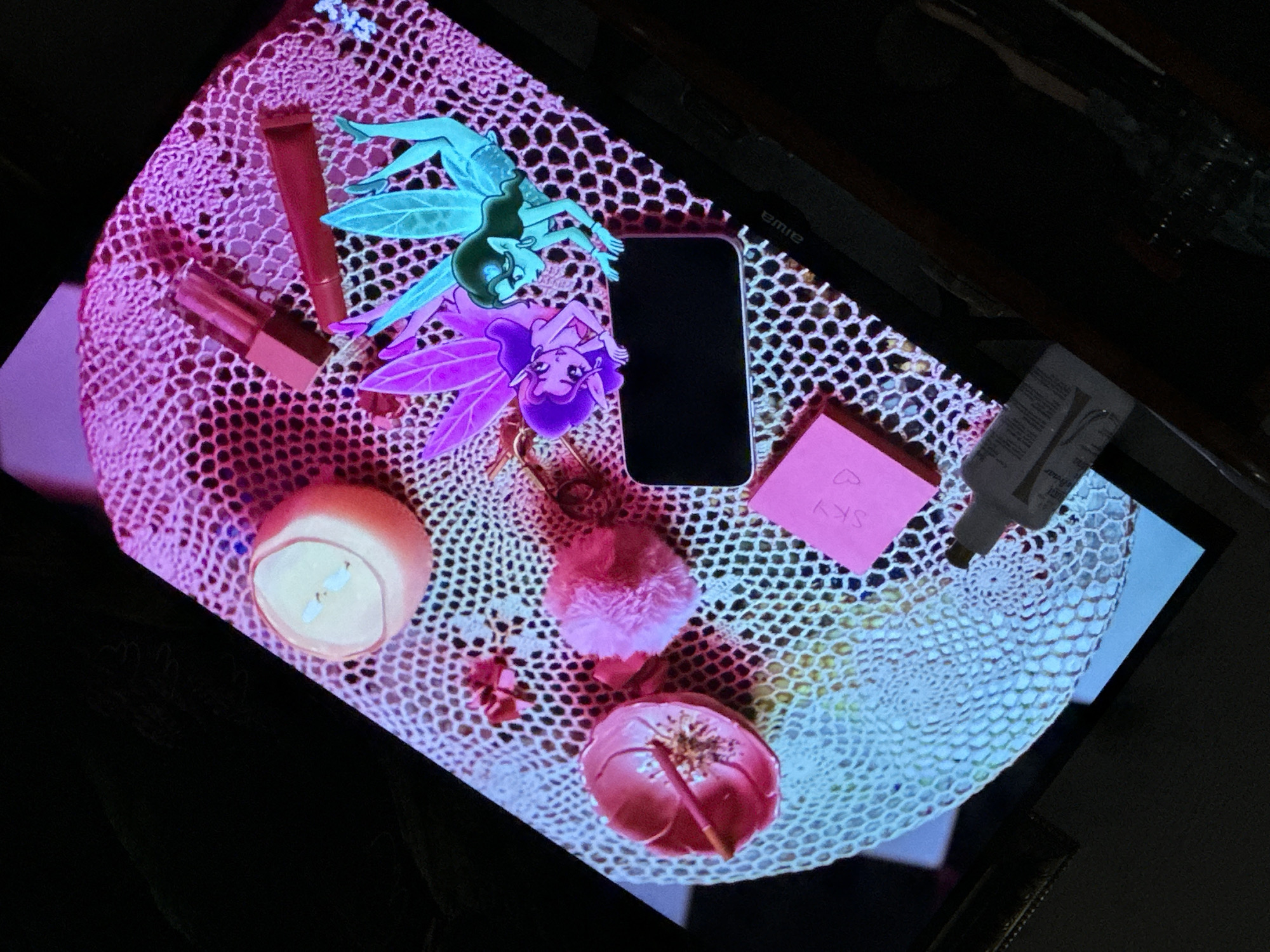 A still-life composition sits on a crocheted, oval-shaped surface, likely a doily, with a hexagonal mesh pattern. The items on the doily include: two lip gloss tubes, one lying horizontally and the other vertically; two cartoon fairy figures, one light blue and one purple; a dark, rectangular mobile phone; a pink square object with "SKY" written on it; a grey bottle with white lettering; a small, round candle in a pink holder; a small, pink pom-pom; and a pink flower-shaped dish containing a brown object. The doily is positioned on a dark surface, possibly a table. The image is bathed in a mix of warm and cool light, creating a purple hue on one side and a cooler, bluer hue on the other. The composition appears to be situated within a residential indoor environment. There is no indication this is in the city of Parroquia Ocumare, Venezuela.