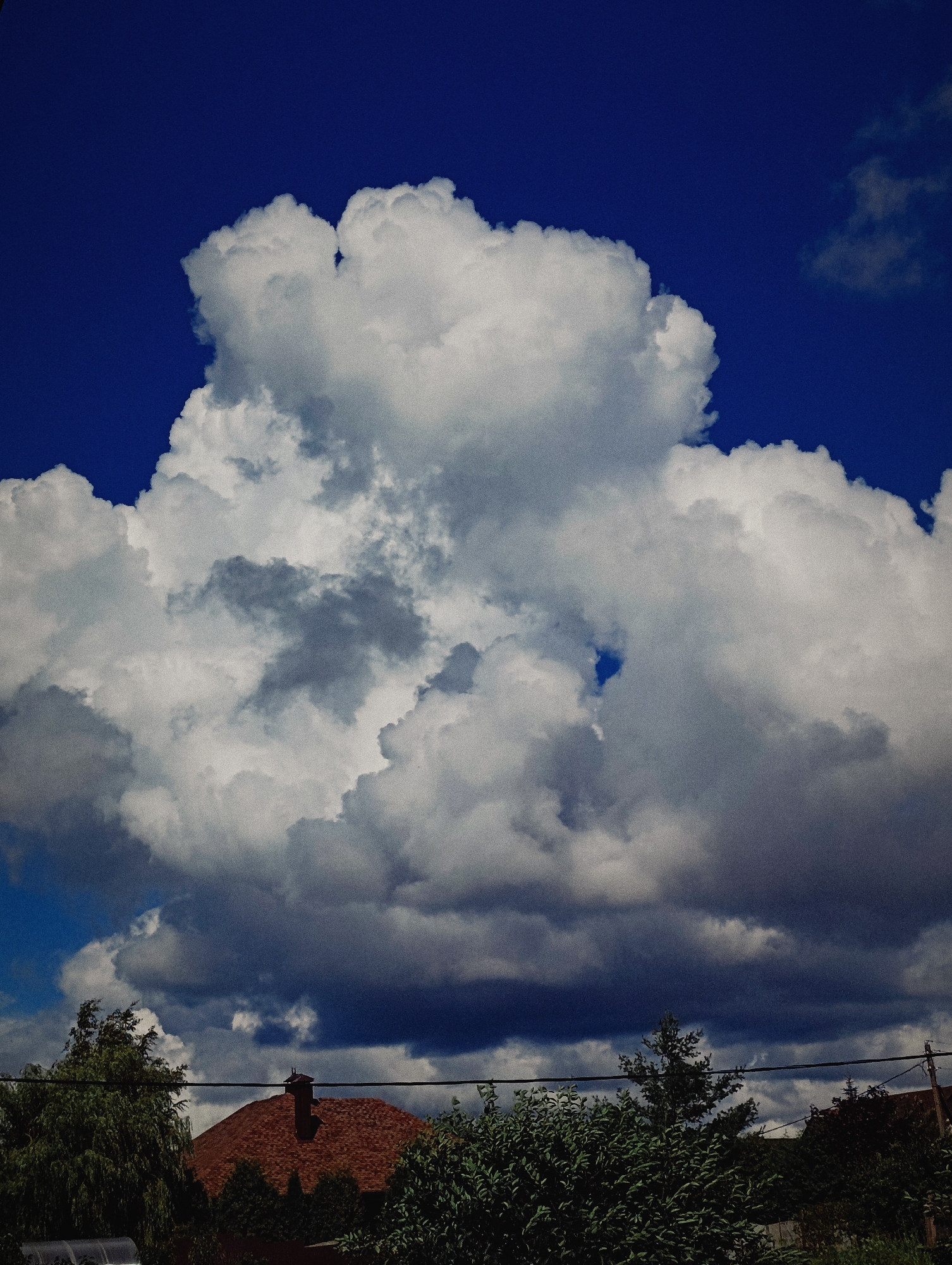 The image shows a dramatic sky with large, fluffy white clouds against a deep blue sky. In the foreground, there are the tops of trees and a portion of a red-tiled roof, suggesting a residential area. Power lines are visible crossing the scene horizontally. The scene is likely a snapshot of the sky over the outskirts of Minsk, Belarus, possibly during a summer afternoon with developing cumulus clouds. There are no people or specific actions depicted. The overall impression is one of natural beauty and a slightly overcast or impending weather scenario.
