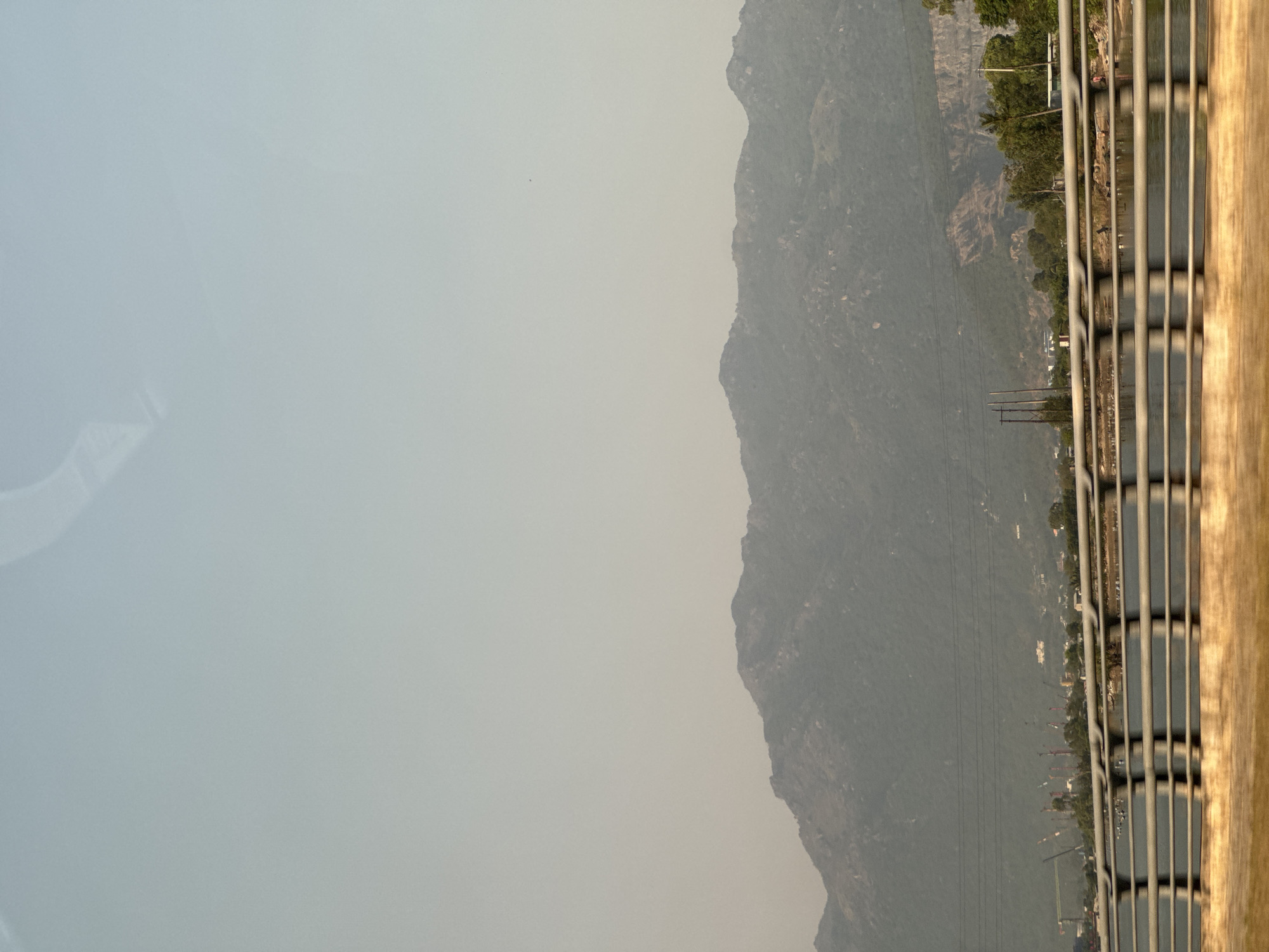 A landscape scene captured from an elevated perspective in Unknown, Unknown shows mountains in the background under a hazy sky. In the foreground, a safety railing is visible. Power lines stretch across the midground landscape. The light suggests it might be either morning or late afternoon.