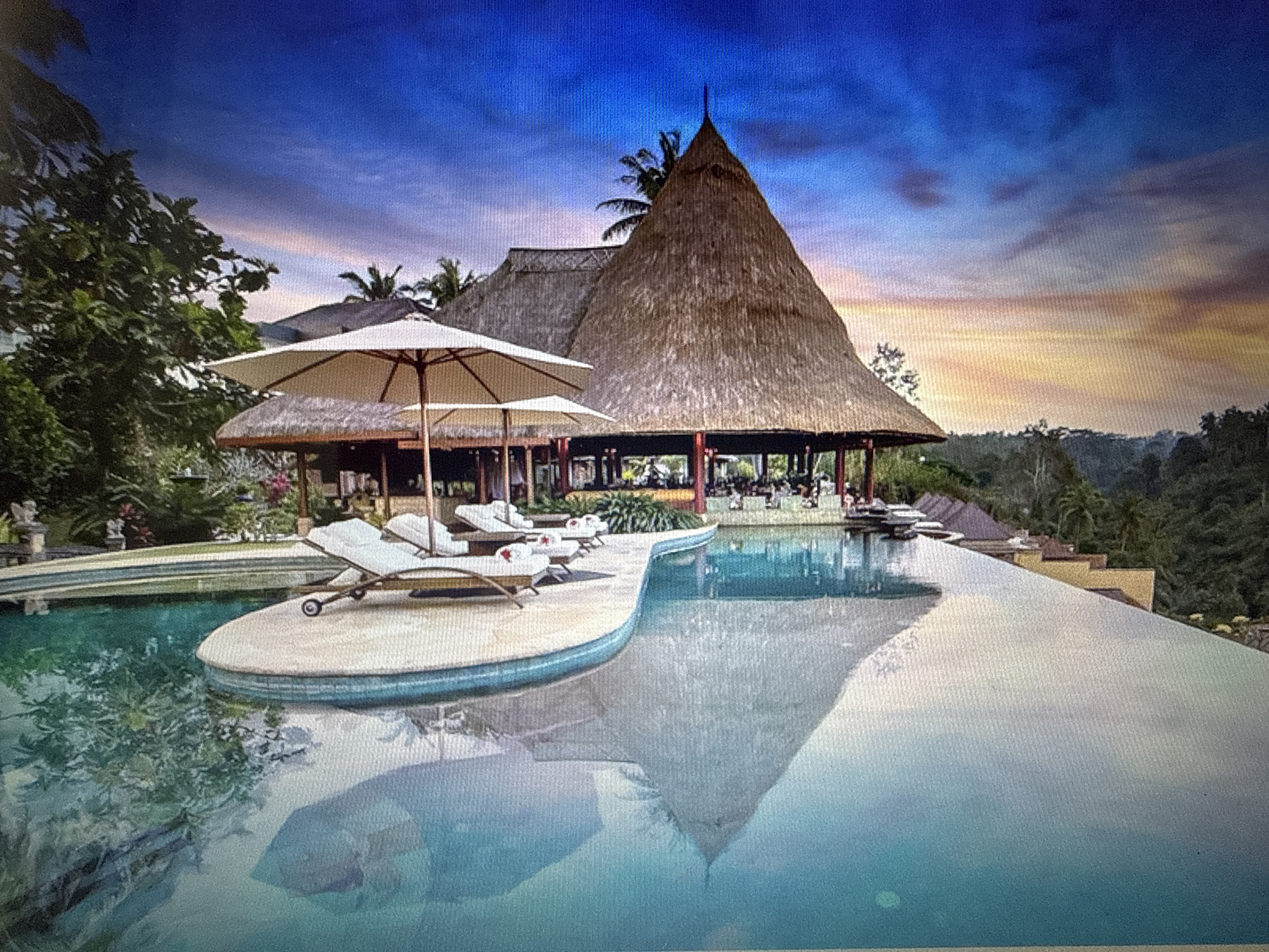 The image depicts a serene infinity pool at what appears to be a resort, overlooking a lush, green landscape. The pool itself is a prominent feature, with clear turquoise water reflecting the sky and surrounding structures. In the foreground, two white lounge chairs with a beach umbrella are positioned on a curved deck extending into the pool.
Behind the pool, a large, thatched-roof building with a conical shape dominates the scene. This building appears to house an open-air dining or lounging area, with tables and chairs visible. Additional structures with similar thatched roofs are visible further back, nestled amongst the trees. The architecture suggests a tropical or exotic location.
The sky above is a vibrant gradient of deep blue at the top, transitioning to hues of orange and yellow near the horizon, indicative of either sunrise or sunset. Silhouetted palm trees and other foliage frame the scene, adding to the tropical ambiance. The overall impression is one of tranquility and luxury.
Regarding the location in the city of Pushkino, Russia, there is no visual evidence within the image to support this claim. The environment and architecture are not characteristic of Pushkino or typical Russian landscapes. The scene strongly suggests a location in a tropical or subtropical region.