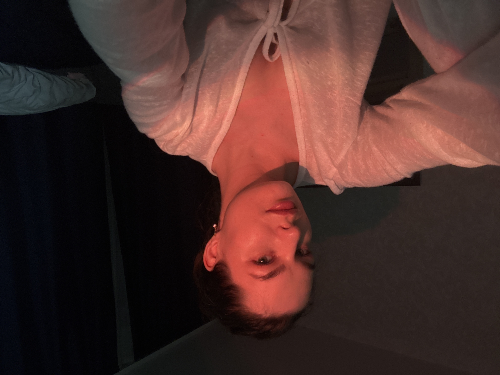 A young woman with her hair pulled back is seen from the chest up, with a red light illuminating her face and upper body. She is wearing a white, open-fronted top tied at the chest with a bow. Her left arm is bent, and her hand is resting on what appears to be a bed covered with light-colored bedding, partially visible to the right. The background is dark, with a hint of a wall texture on the left. The image was taken in a bedroom environment in Chaikovsky, Russia.