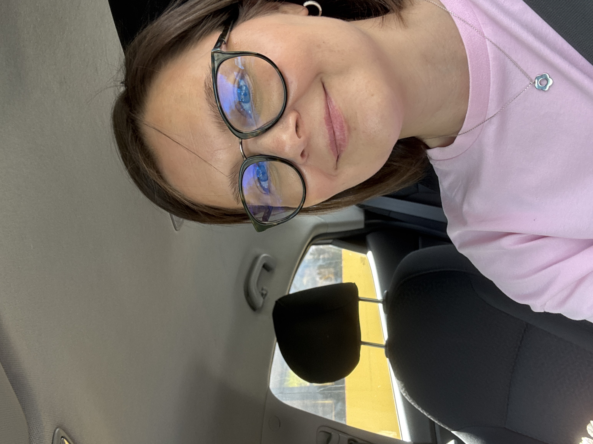A medium shot shows a young woman with short brown hair and glasses sitting in the passenger seat of a car. She is wearing a pink t-shirt and a silver necklace with a flower pendant. The background shows the interior of the car, including a headrest and a window with a yellow object visible outside. The image does not provide any specific details about the location in the city of Izhevsk, Russia.