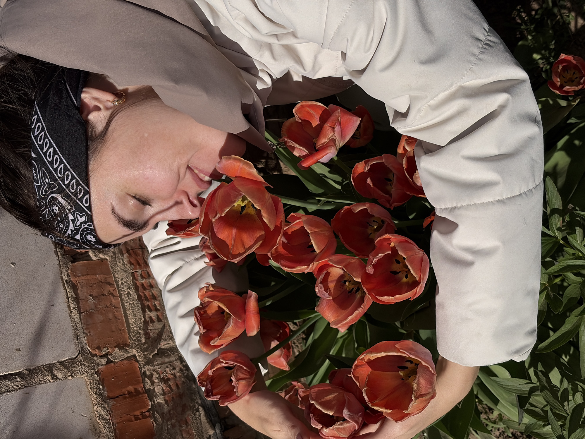 A person lies on their back surrounded by a bouquet of orange tulips. The person is fair-skinned, wearing a black bandana on their head, a gold earring, and a light-colored jacket. The tulips are in full bloom, with deep orange petals and dark centers. The person's hands hold the base of the bouquet. The background includes brickwork and green foliage. There is nothing suggesting that the location is Nizhnii Vishur, Russia.
