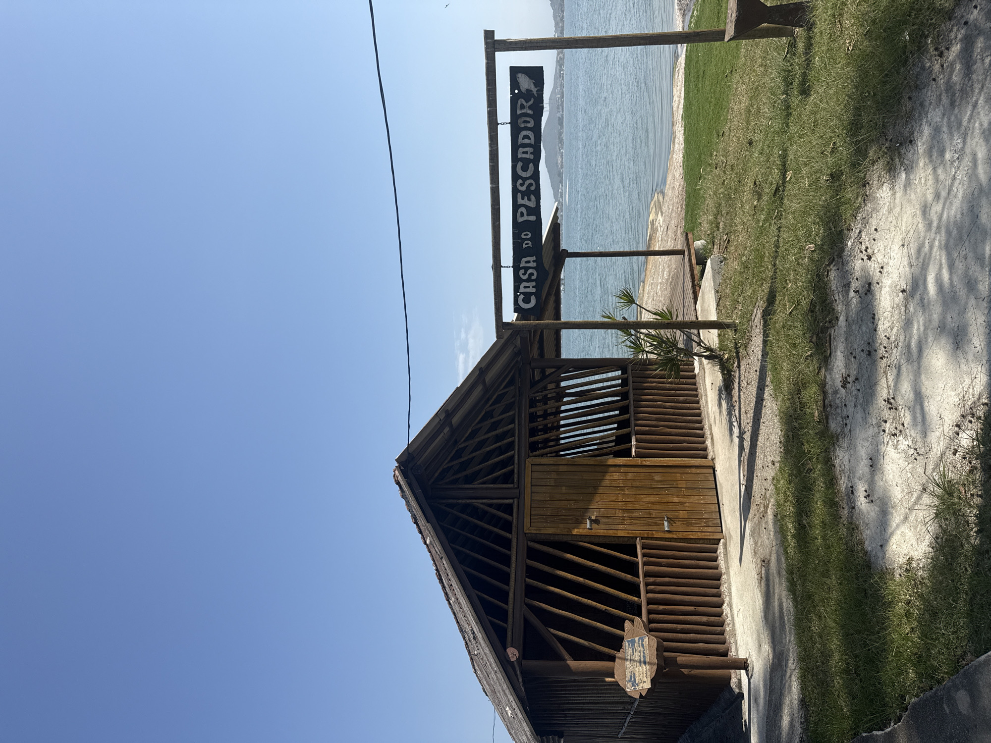 An outdoor scene in Iguaba Grande, Brazil, features a rustic wooden structure with a thatched roof, possibly a fisherman's hut, situated near the water. A signpost in front of the structure reads "CASA DO PESCADOR" in white lettering with a fish graphic on the right. The hut has a wooden door and open sides with vertical wooden slats. The area is paved with concrete in some parts and covered with grass in others. The water body in the background suggests it is a lake or bay, with a distant hilly landscape visible. The sky is clear and blue. There are no people or animals visible in the image. The lighting suggests it is daytime.
