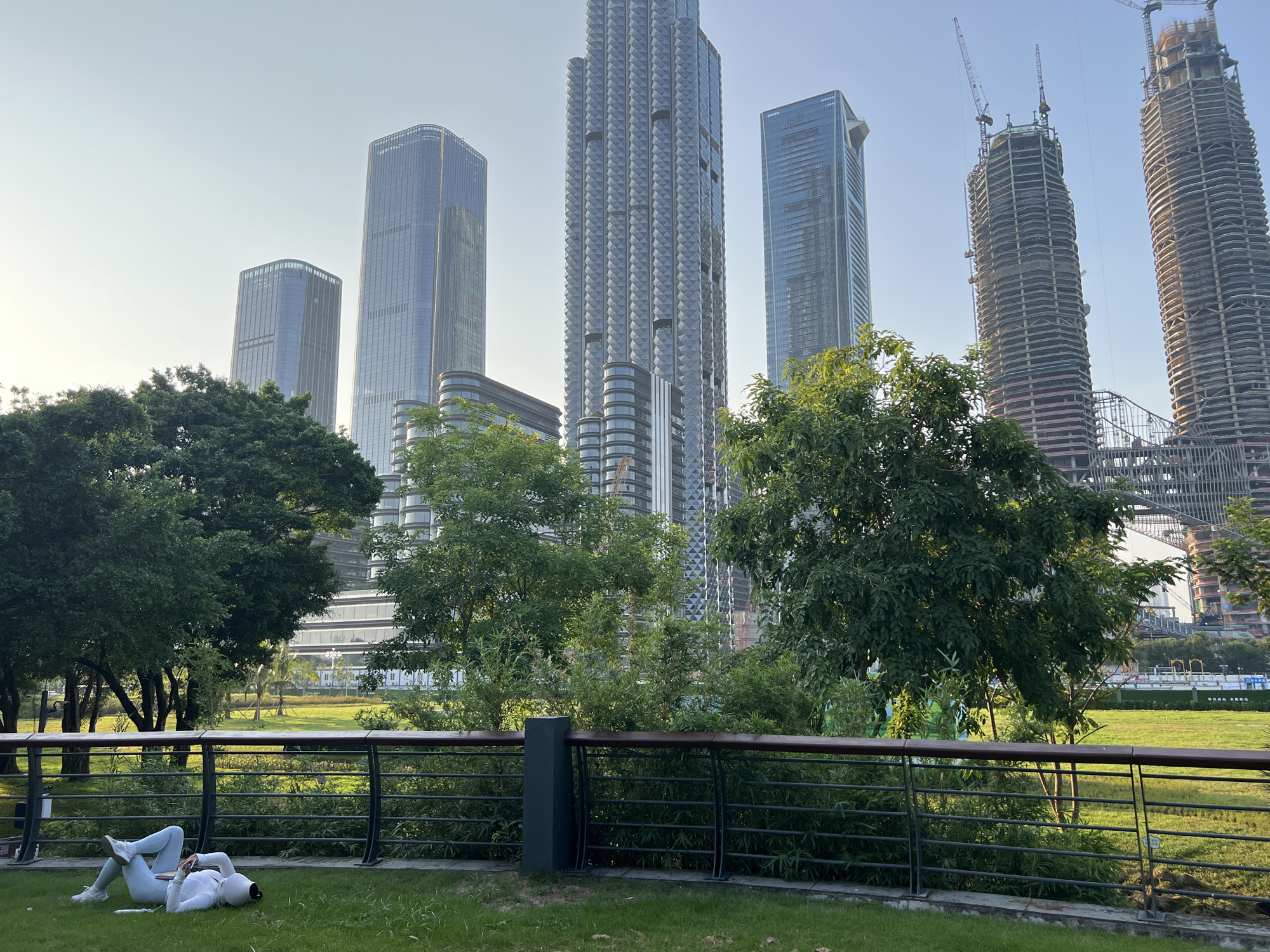 In Nanshan District, China, a person lies on a grassy area, propped up on their elbow and looking at a phone. They are wearing a white long-sleeved shirt and white leggings, with white sneakers. The person is positioned in the foreground, near a dark metal railing with a wooden top. Behind the railing, a lush green lawn stretches towards a cluster of trees. Beyond the trees, a cityscape of modern skyscrapers dominates the background. Some of these buildings are complete, while others appear to be under construction, with visible cranes and exposed frameworks. The sky is clear and a pale blue, suggesting it is daytime. The overall scene depicts a blend of urban development and green space.
