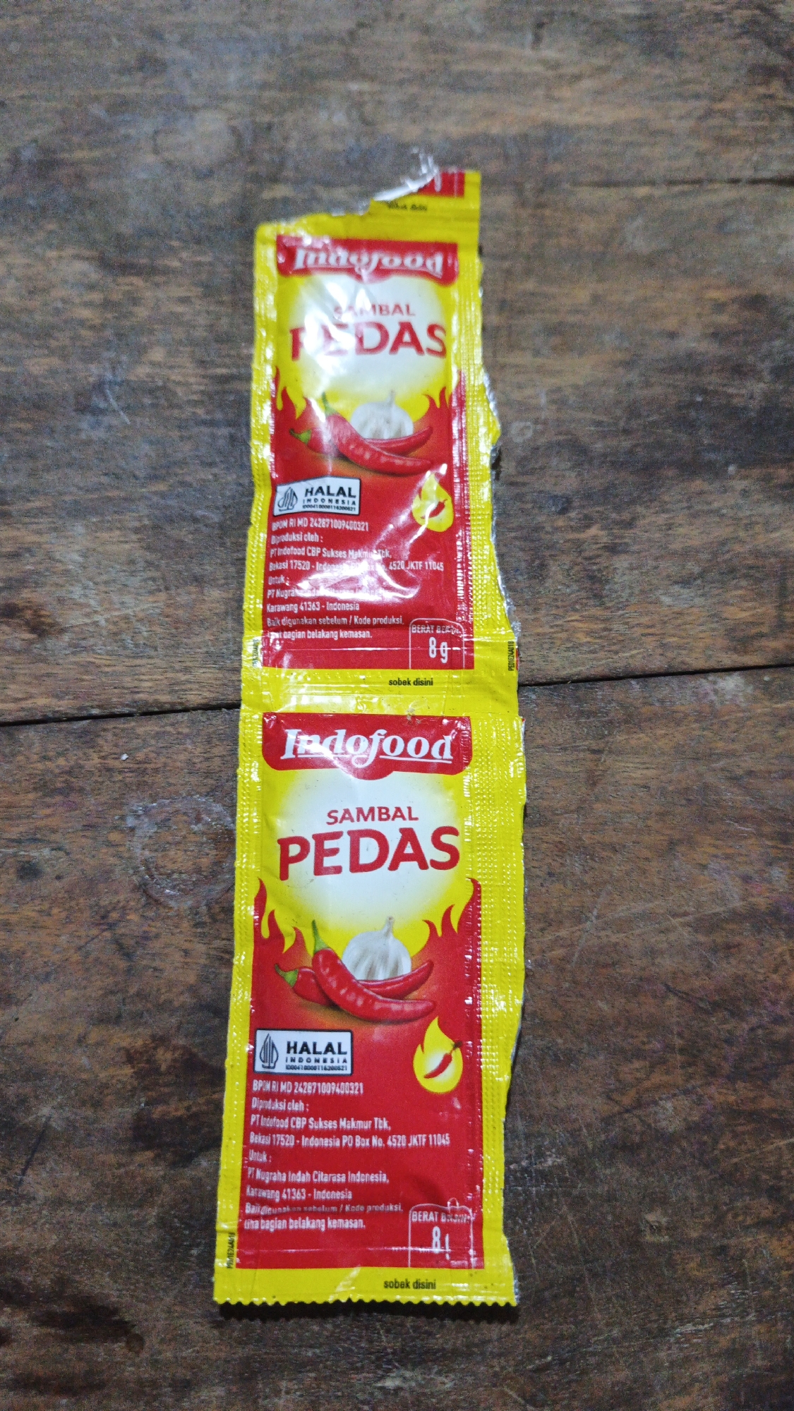 The image shows two packets of Indofood Sambal Pedas, a chili sauce, against a wooden background. Both packets are predominantly yellow and red, with the Indofood logo and the product name "SAMBAL PEDAS" prominently displayed. The top packet is slightly torn at the top. Below the product name, there are images of chili peppers and garlic. Both packets also feature a HALAL Indonesia logo and production information. One packet has "BERAT BERSIH 8g" written on it. The setting appears to be outdoors or in a location with natural wood textures. There are no people or animals in the image.