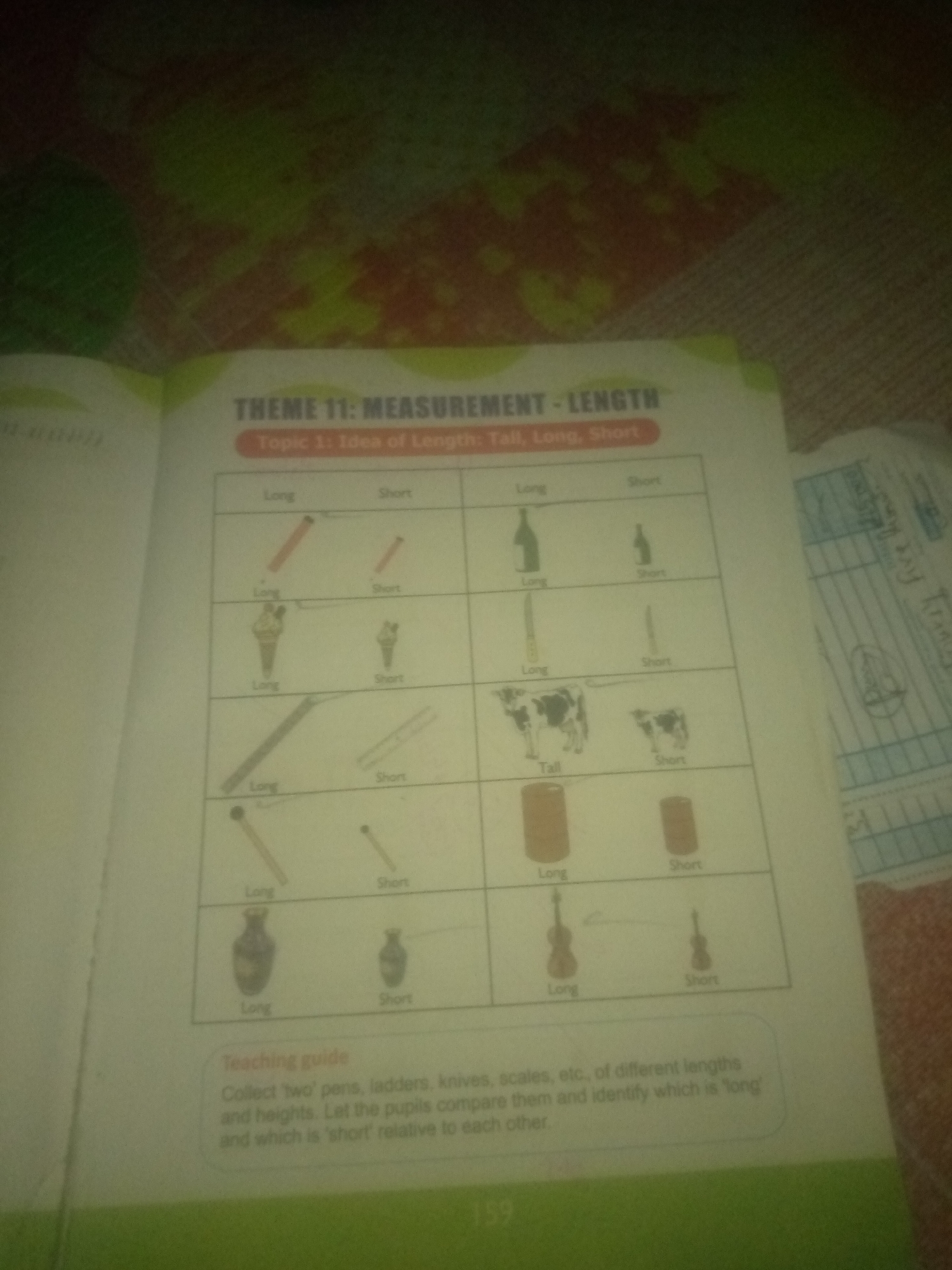 The image is a photograph of a page from a textbook or workbook. The page is titled "THEME 11: MEASUREMENT - LENGTH" and "Topic 1: Idea of Length: Tall, Long, Short". Below the title is a table with images illustrating the concepts of long and short. The table contains rows with pairs of objects, each labeled as either "Long" or "Short". The objects depicted include pencils, ice cream cones, bottles, straws, cows, cans, matchsticks, and vases. A "Teaching guide" section at the bottom of the page provides instructions for educators. The image itself appears to be taken in a room with a patterned textile in the background, possibly a carpet or upholstery. The lighting is dim, and the focus is not sharp. There is no identifiable location in Jauro Yinu, Nigeria.