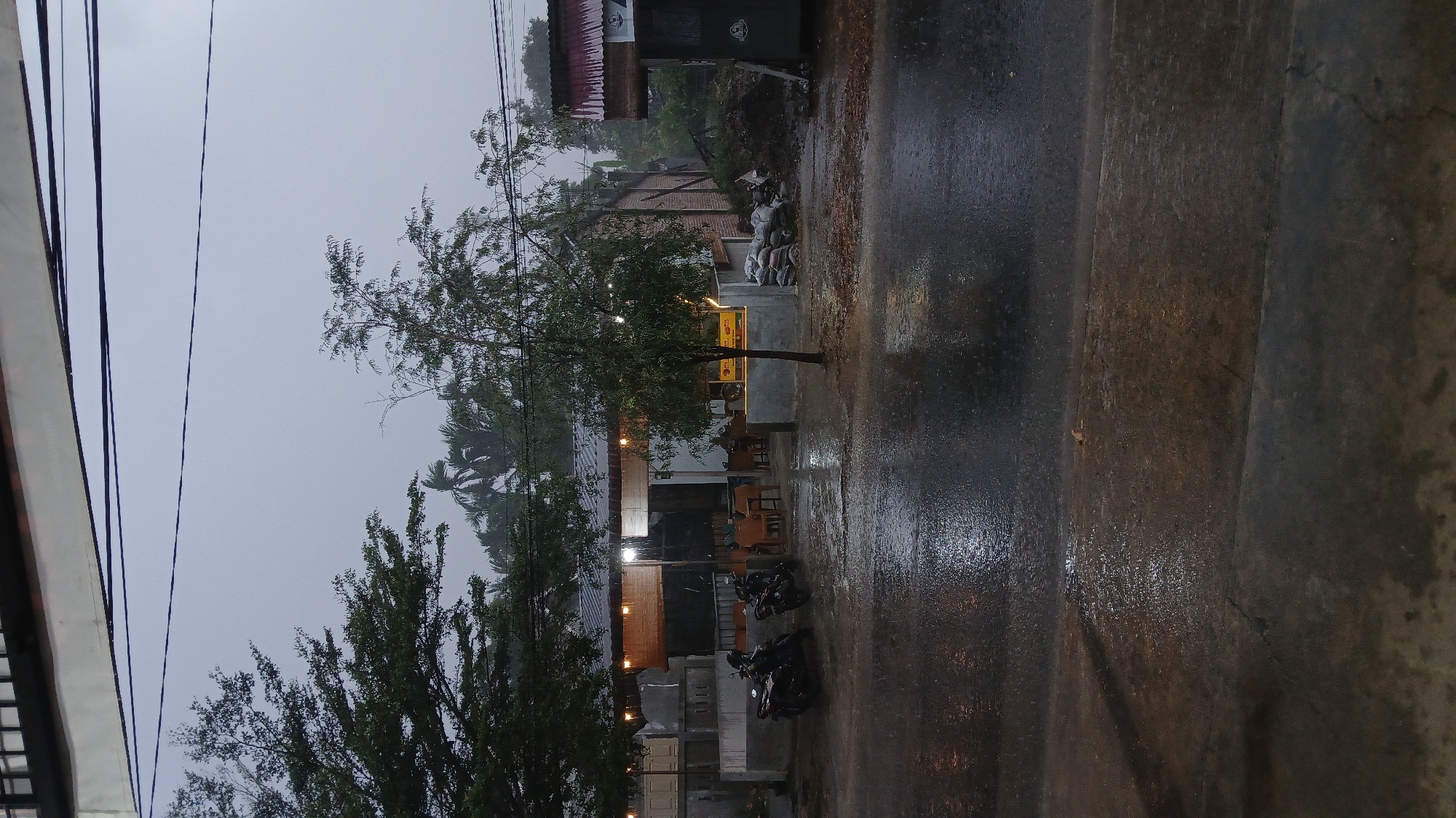 The image shows a rainy day in Lhokseumawe, Indonesia. A street is wet from the rain, reflecting the sky. On the left, a building with visible electrical wires extends from the top to near the bottom of the image. A tree grows on the left side of the building. Across the street, there is a building with its light on. Several parked motorbikes are visible in front of the building. Further back and to the right are brick buildings and what appears to be a stack of bags. The sky is overcast and the overall scene is dimly lit.