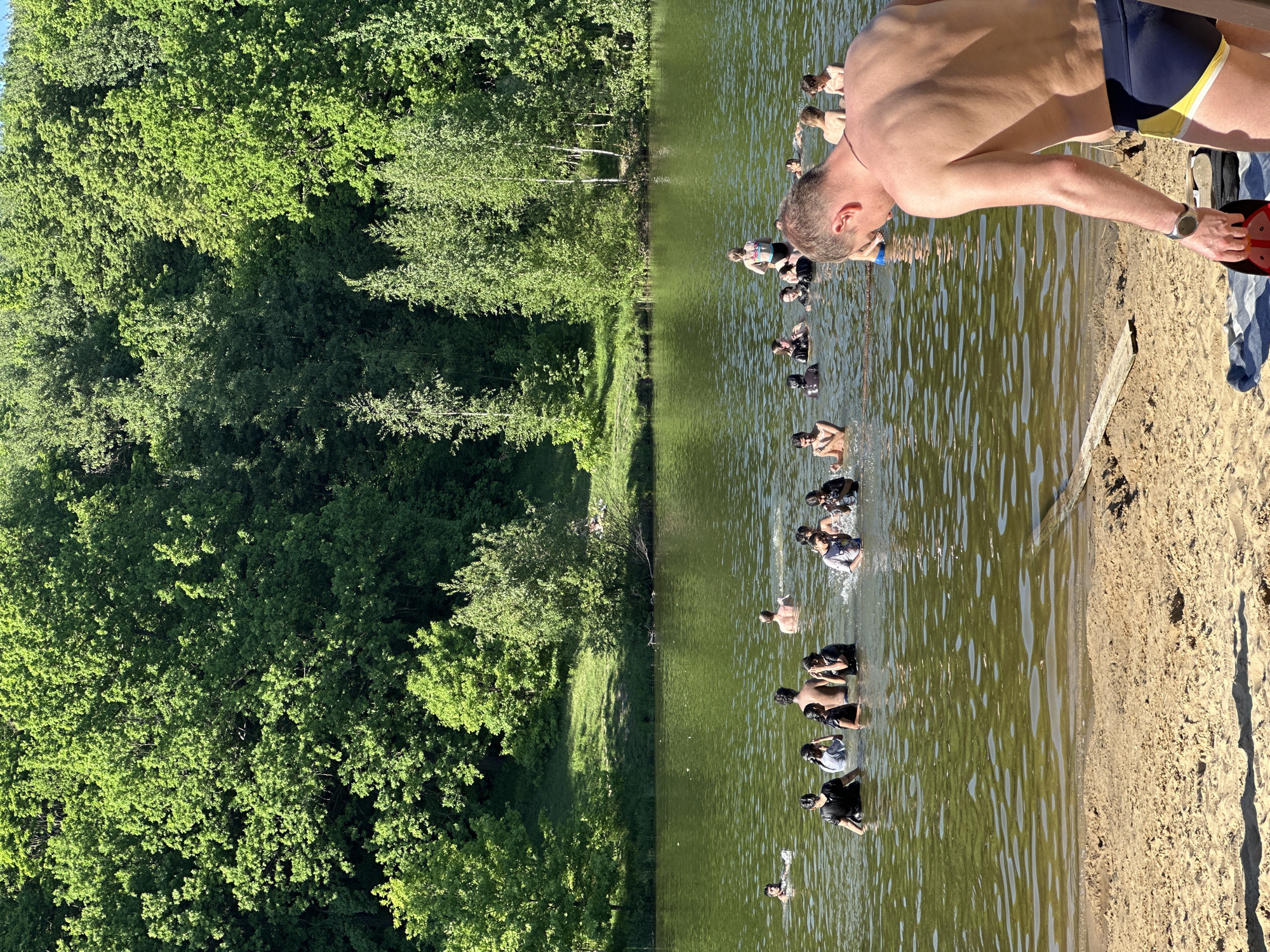 A group of people are in the water at a sandy lakeside location with trees in Кузнечиха, Russia. A man with a wristwatch, wearing blue and yellow swimming trunks, stands on the sand beside shoes, a red object, and a striped towel. He appears to be looking at the people in the water.