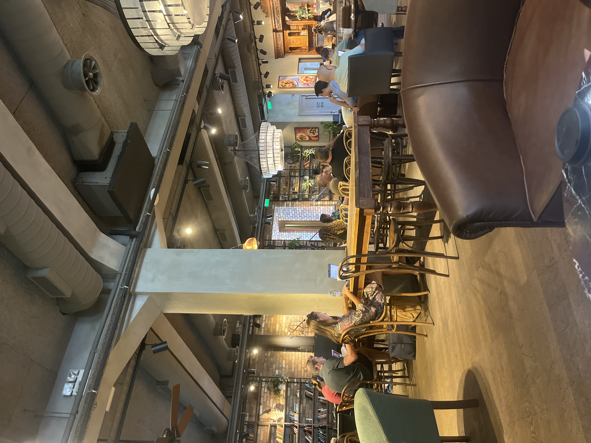 The image depicts the interior of a cafe, likely in Agios Athanasios Municipality, Cyprus, with a warm and inviting atmosphere. A large communal wooden table dominates the center of the frame, around which several patrons are seated, engaged in various activities. A woman in a patterned dress is typing on a laptop, while a man in a dark green polo shirt sits beside her, also with a laptop open. Another person with headphones is visible further down the table. The cafe features exposed brick walls adorned with bookshelves filled with books, creating a rustic charm. Industrial-style ventilation ducts and lighting fixtures are visible on the ceiling, contrasting with the softer elements of the decor. In the background, more seating areas are visible, including comfortable leather armchairs and other tables occupied by patrons. The lighting is a mix of ambient and focused lights, casting warm glows and shadows throughout the space. The overall impression is that of a lively and functional space for work, study, and socializing.
