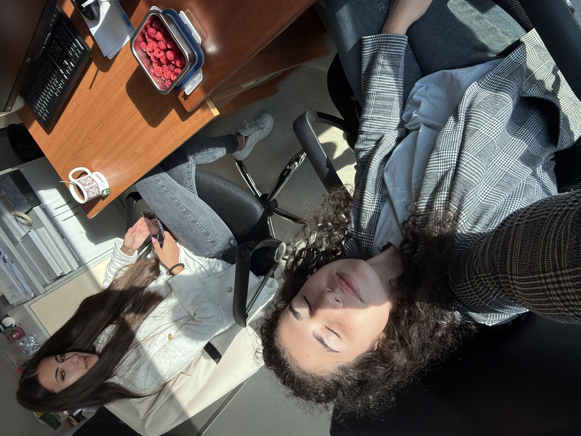 The image depicts two young women in an office environment, likely located in Sofia, Bulgaria. The woman in the foreground is lying back in an office chair, with her eyes closed and sunlight illuminating her face. She is wearing a gray and white plaid blazer over a light blue shirt and dark denim jeans. Her curly, dark hair is spread around her.
The second woman is seated in another office chair, facing slightly away from the camera, towards a desk. She has long, dark hair and is wearing a white textured jacket over a dark top and jeans. She is holding a mobile phone in her right hand and looking towards it.
On the wooden desk, a white mug with a folk art design and the inscription "MAPEX" is visible, with a spoon inside. Next to the mug is a metallic container filled with raspberries. A computer keyboard and monitor are also on the desk, suggesting a typical office workspace. The overall lighting suggests it is daytime, with strong sunlight casting shadows. The presence of office furniture and equipment indicates a professional setting.