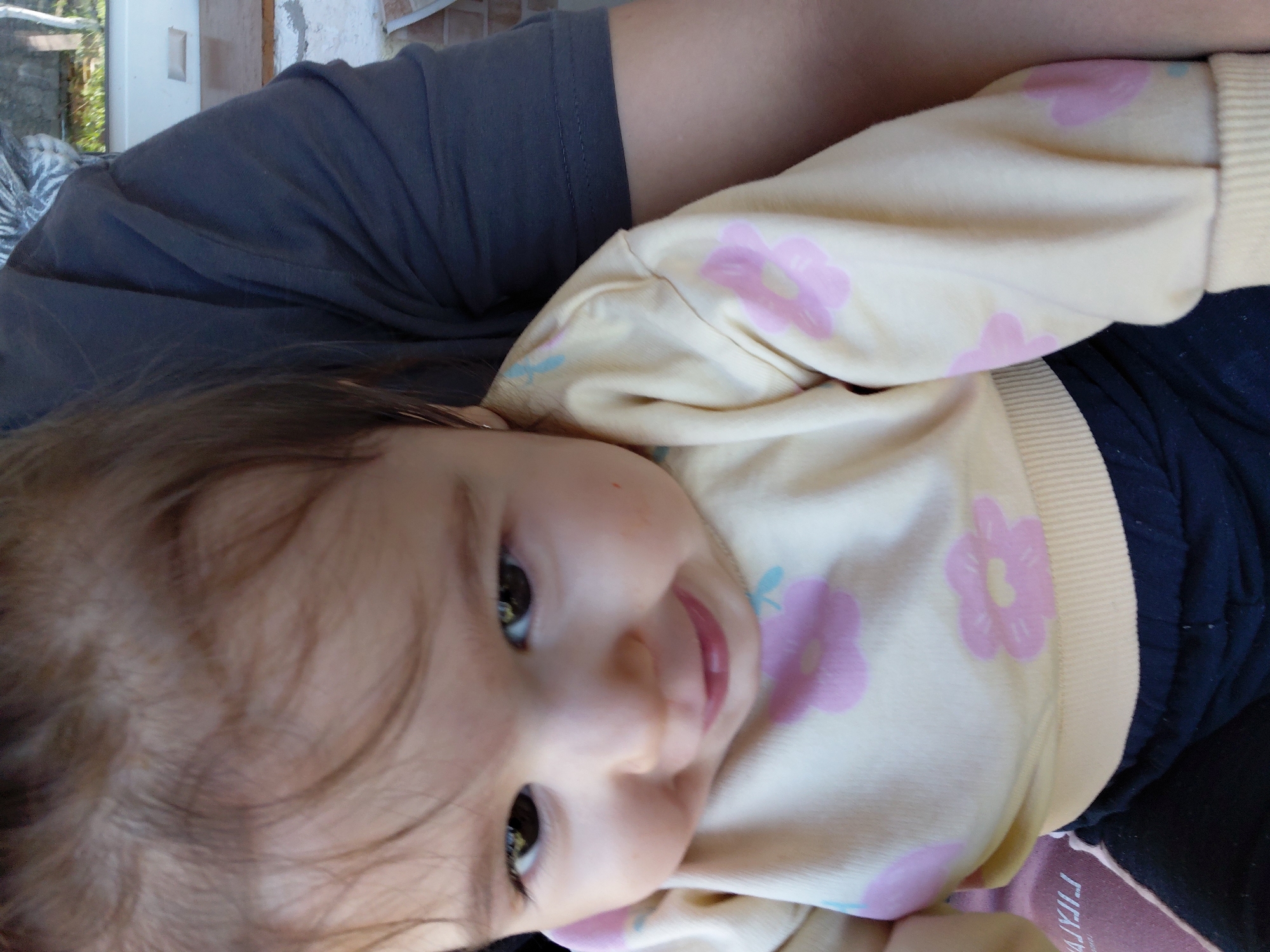 A baby is resting in someone's arms. The baby is wearing a light yellow shirt with pink flower patterns and dark pants. The person holding the baby is wearing a gray shirt. The photo was taken indoors. The exact location in the city of Samovodene, Bulgaria cannot be determined from the provided image.