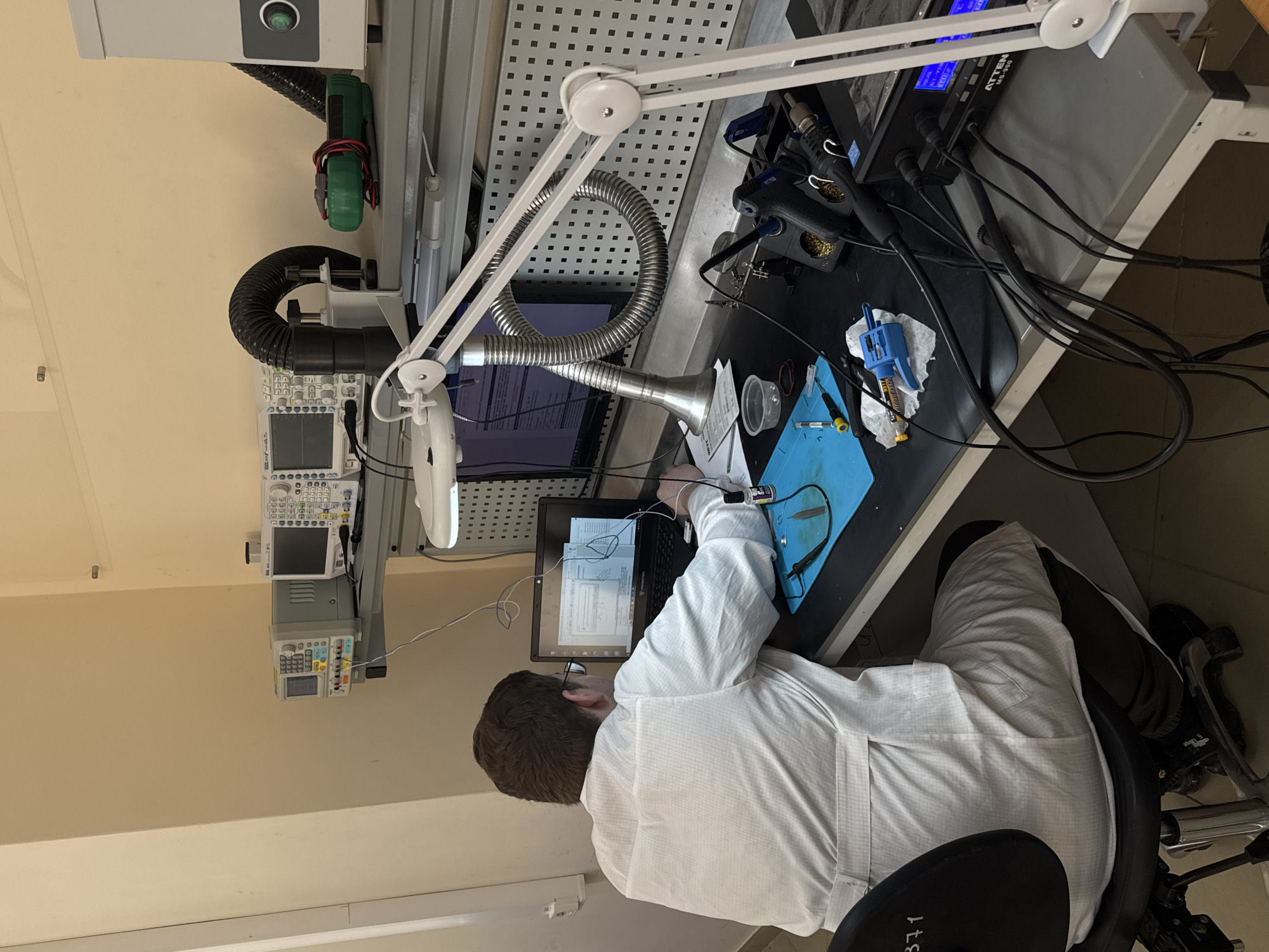 A person wearing a white lab coat is seated at a workbench in what appears to be an electronics or engineering laboratory. The individual is working on a laptop, with various electronic components and tools scattered across the desk. An arm-mounted magnifying lamp is positioned to provide illumination and magnification for detail work. Electronic testing equipment is visible in the background, including oscilloscopes and a soldering station. The location is potentially Perm, Russia, though that is not verifiable from the image itself.