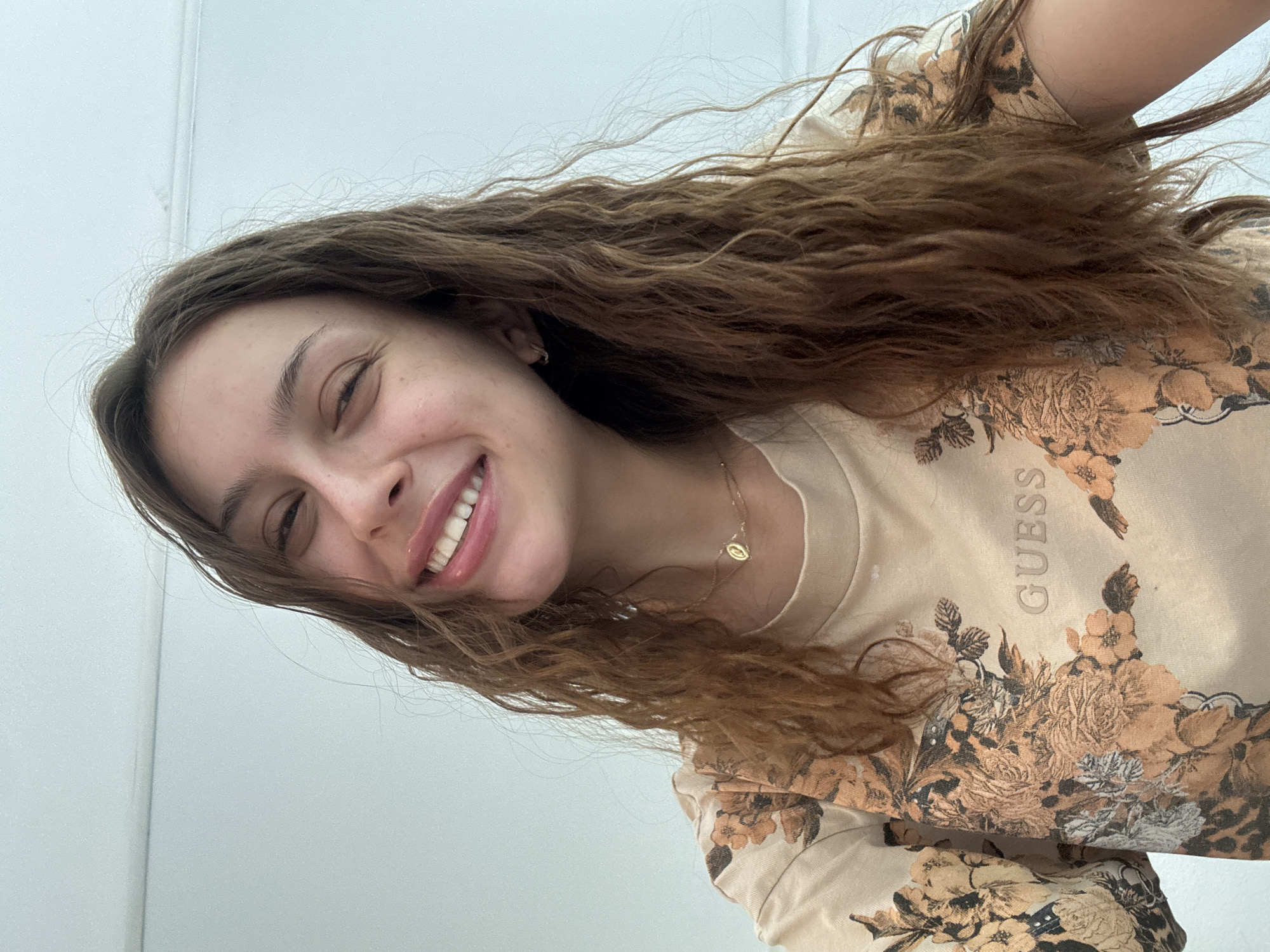 A young woman with wavy brown hair and light skin is smiling directly at the camera. She is wearing a beige shirt with floral designs and the word "Guess" printed on it. A delicate gold necklace with a small circular pendant is visible around her neck. The background is a plain, light-colored wall. The image does not suggest or contain any identifiable information relating to Milan, Italy.