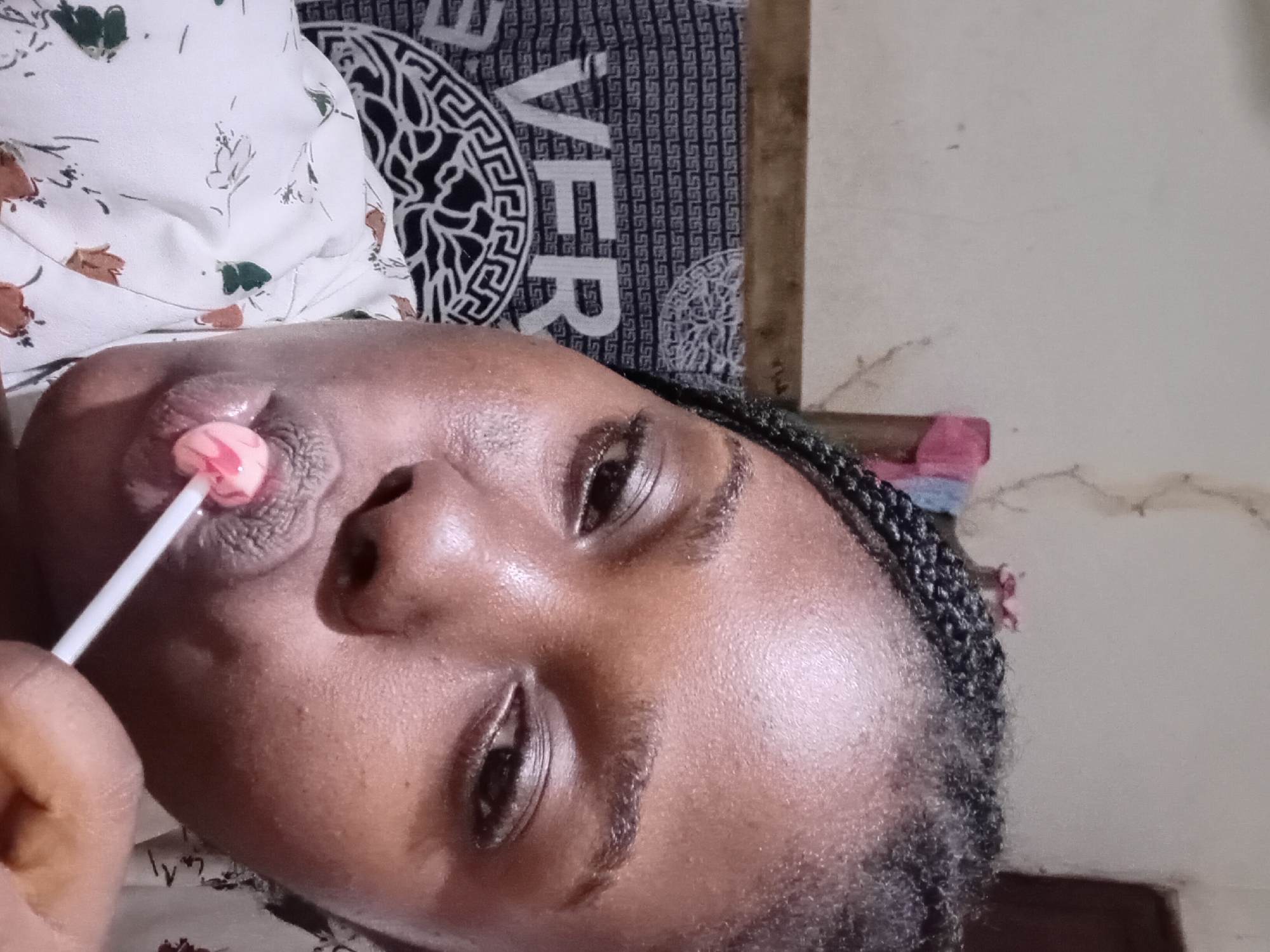A close-up, eye-level shot shows a Black female with dark skin and braided hair, indoors in Ejule, Nigeria. She is holding a pink lollipop to her mouth, appearing to be sucking on it. She is wearing a white shirt with a floral pattern. Behind her, a dark patterned fabric with the word "VERS" visible is draped. The background wall is textured and lightly colored.