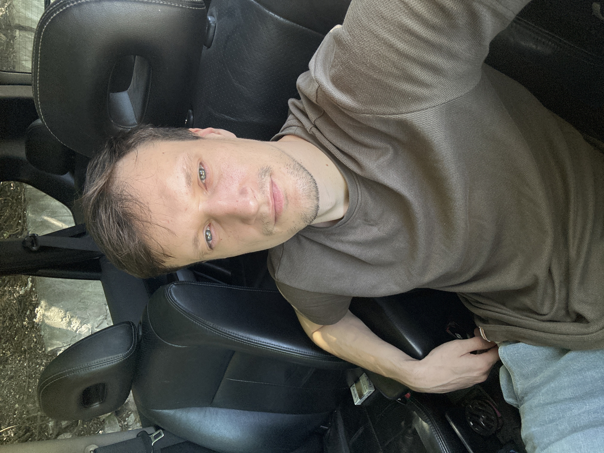 A man is seated in the driver's seat of a car, facing the camera. He has short, light brown hair and is wearing a dark brown t-shirt and light blue pants. His right arm is resting on the center console, and his hand is partially visible. The interior of the car appears to be black leather. Outside the car, sunlight is visible, indicating it is daytime. The location is not specified in the image, and there is no information to suggest it is in Makiv, Ukraine.