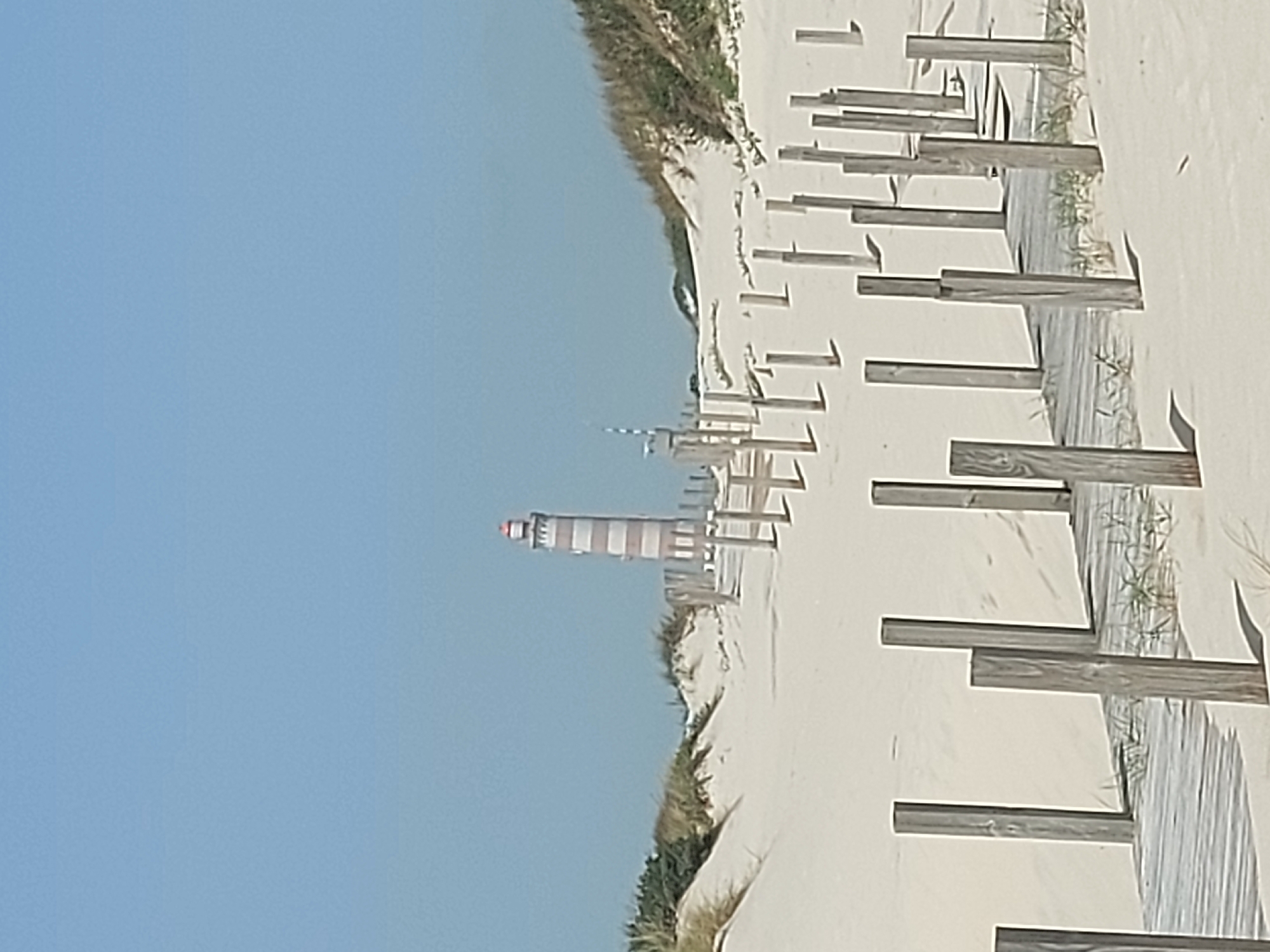 The image shows a beach scene in Gafanha da Nazaré, Portugal. A striped lighthouse stands in the distance, visible against a clear blue sky. The foreground features a sandy beach with wooden boardwalk-style barriers lining the path. Vegetation grows on the dunes separating the boardwalk from the open sand. There are no people visible in the image.