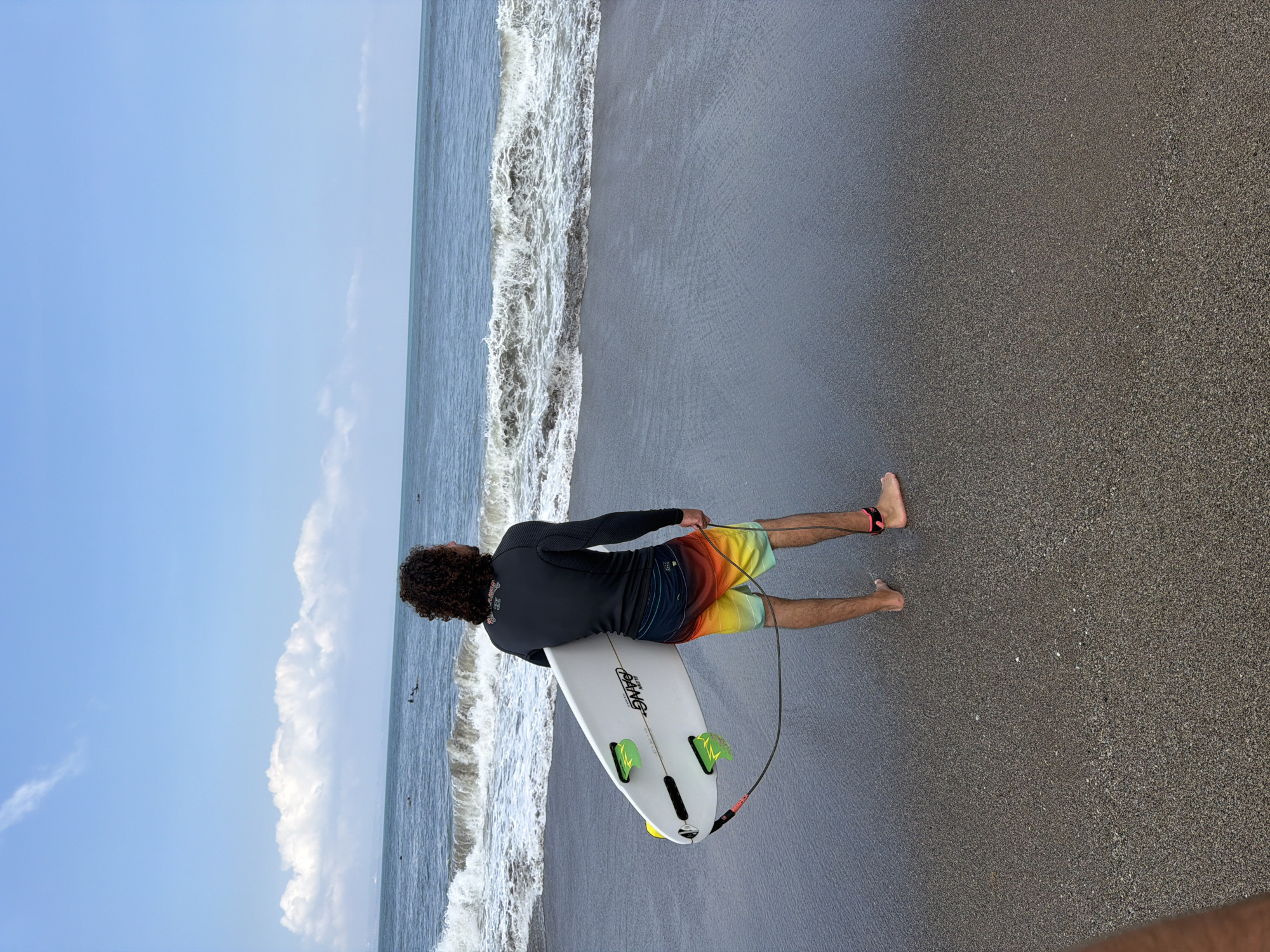 A surfer with curly hair stands on a dark sand beach, facing the ocean. He is wearing a black long-sleeve rashguard and colorful, ombre-patterned swim trunks. In his right hand, he holds a white surfboard with green fins and a leash attached to his ankle. The ocean in front of him has white-capped waves and a distant horizon line. The sky is a clear blue with scattered white clouds. The scene is captured from a slightly elevated angle, looking down at the surfer and the beach. The location is identified as the city of North Kuta, Indonesia.