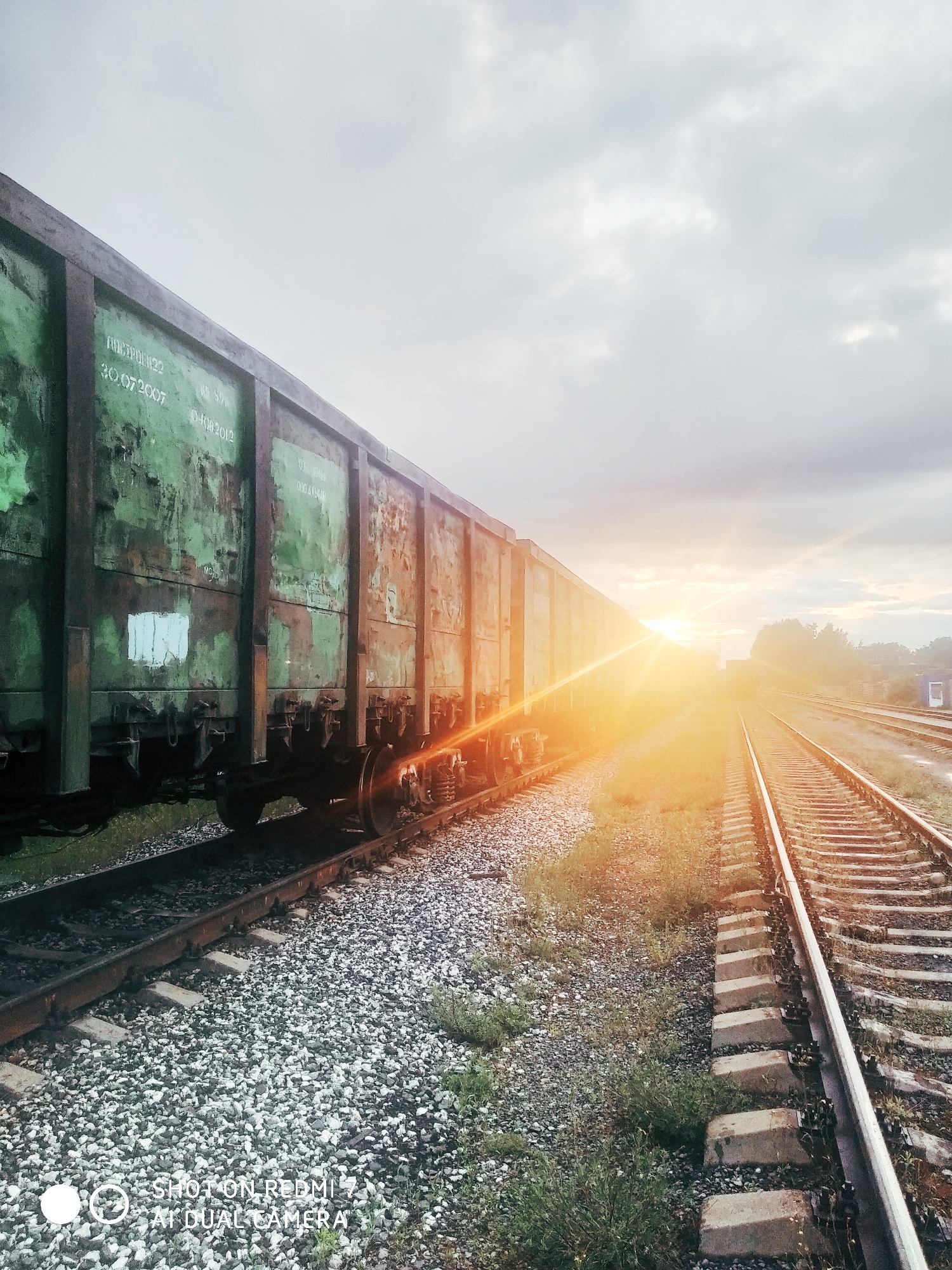 An eye-level shot captures a long, weathered green freight train car on the left, positioned on a railway track. The perspective extends forward and to the right, revealing multiple sets of railway tracks disappearing into the distant horizon under a bright, low-angled sun. A prominent golden sunburst lens flare emanates from the right side of the image, where the sun is setting or rising, casting a warm glow across the scene.
The train car, likely a gondola or hopper car, shows signs of rust and faded paint, indicating age and heavy use. White stenciled text is visible on its side, including "ПОСТРОЕН 22" (Built 22), "30 07 2007" (July 30, 2007), "КР 59%", and "01 08 2012" (August 01, 2012), which likely denote manufacturing and repair dates or codes.
The tracks are composed of metal rails on concrete sleepers, embedded in grey gravel ballast. Patches of green grass and weeds grow between the tracks and along the embankments. In the far distance on the right, blurred foliage from trees is visible, with a faint suggestion of industrial structures or another train. The sky above is overcast with soft, grey clouds, through which the sun pierces.
The image depicts a quiet moment at a rail yard or siding, possibly in Chaikovsky, Russia, during either sunrise or sunset, given the warm, dramatic lighting. The train appears stationary, suggesting a pause in operation. A watermark "SHOT ON REDMI 7 AI DUAL CAMERA" is visible in the bottom-left corner.