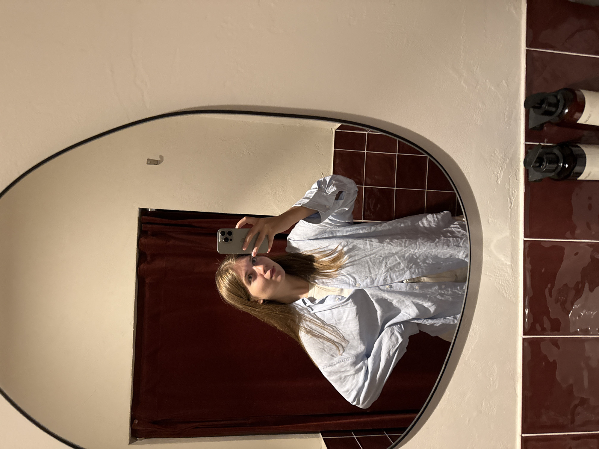 A young woman takes a mirror selfie in what appears to be a bathroom. The mirror is an irregular shape with a black frame, reflecting a view of a burgundy curtain and tile wall. She is wearing a light blue button-down shirt. The bathroom wall around the mirror is off-white and textured. A hook is visible on the wall above the curtain. Bottles are arranged to the right of the mirror, on the tiled wall. The scene does not explicitly reveal a location in Belgrade, Serbia.