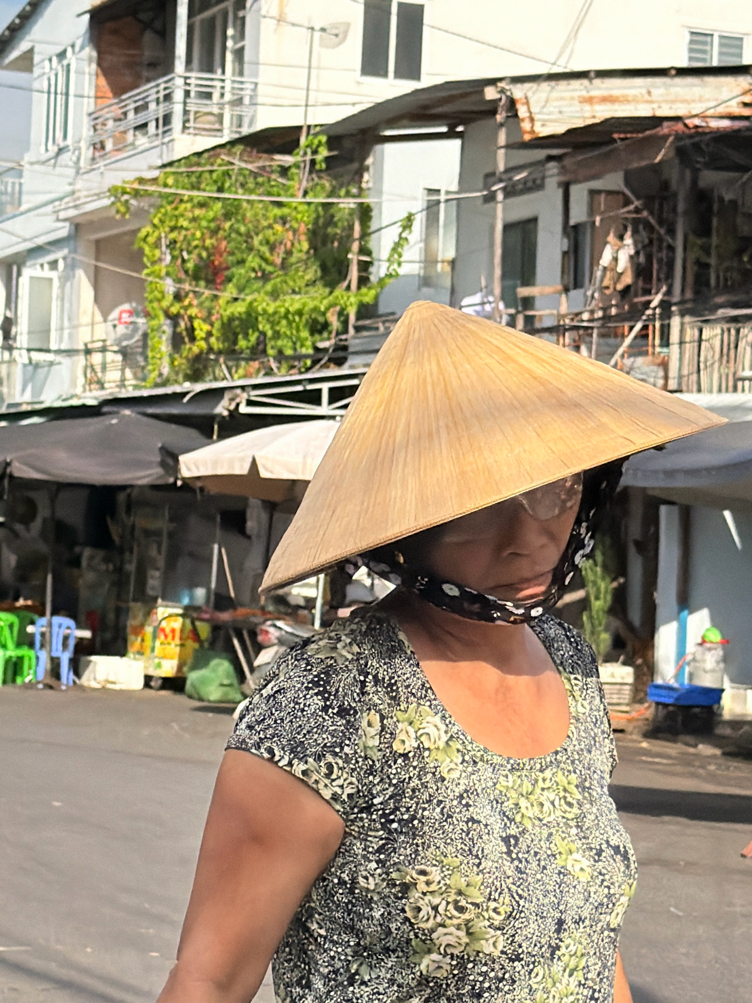 A woman wearing a conical hat and a floral print shirt walks down a street in Nha Trang, Vietnam. The street is paved and appears to be lined with buildings and market stalls. The buildings are a mix of old and new, with some appearing weathered and others more modern. The market stalls are covered by awnings and sell various goods. The woman is walking from left to right in the frame and appears to be looking down. The lighting suggests it is daytime.