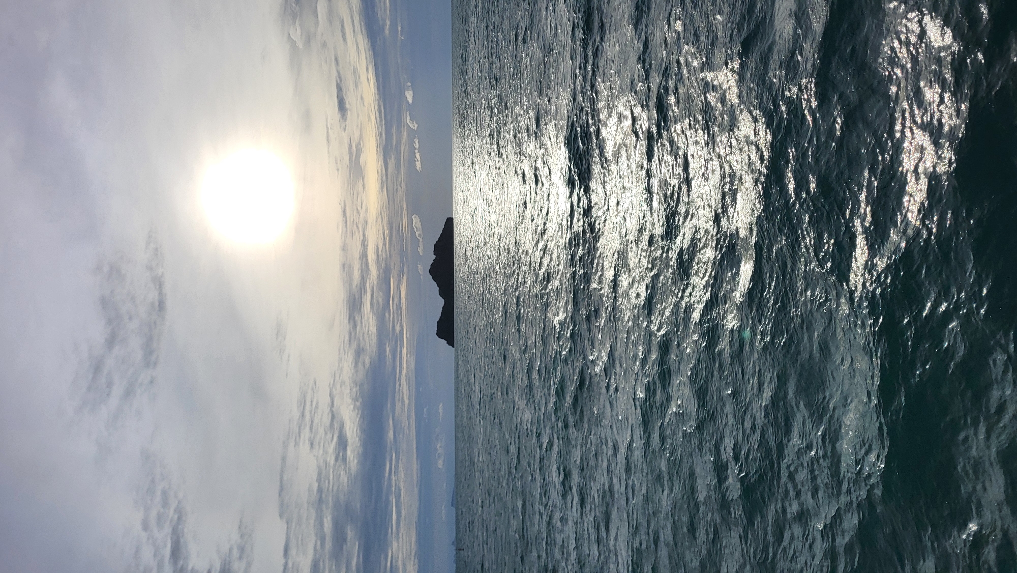 A lone island with a jagged, rocky peak is silhouetted against a bright sun peeking through cloudy skies. The sun's rays cast a shimmering path across the deep blue, choppy waters of the sea. The scene, likely captured from a boat, evokes a sense of peaceful isolation and natural beauty, characteristic of the waters surrounding Ko Pha-ngan District in Thailand.