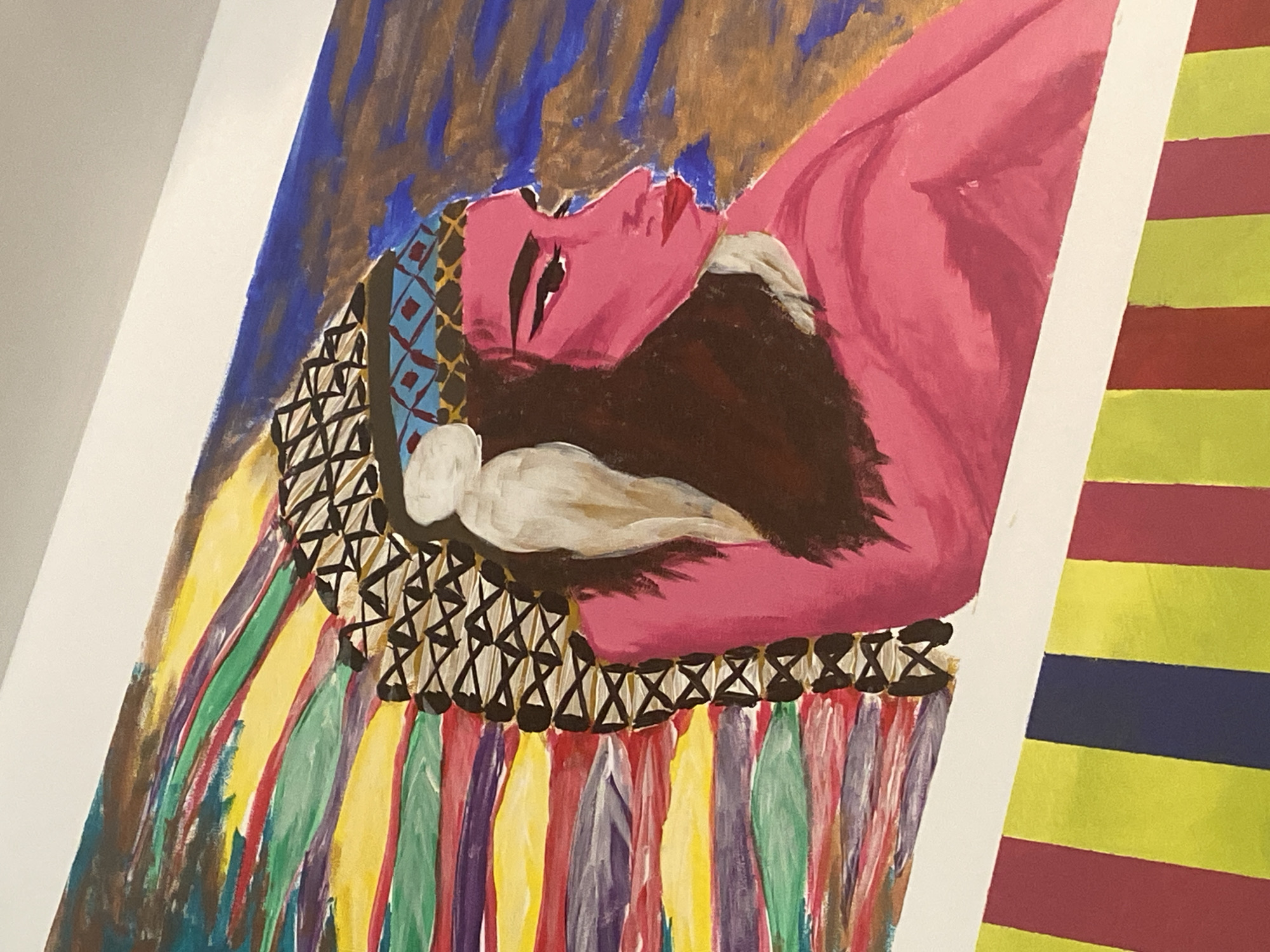 This media file presents a close-up view of a vibrant, stylized painting, likely displayed in an indoor setting on a white surface. The main subject of the painting is a reclining figure, possibly a woman, depicted with striking pink skin and dark, flowing hair.
The figure's face shows closed or heavily shadowed eyes and slightly parted lips, suggesting a state of repose, sleep, or deep thought. She is adorned with an elaborate, colorful garment or headpiece. The upper part of this attire features intricate patterns, including blue and red diamond shapes near her head and a band of black crisscross designs around her neck and shoulders. Below this, the garment flows downwards into broad, vertical stripes in a rich palette of colors, including yellow, green, red, purple, and hints of blue.
The background of the painting behind the figure consists of textured brushstrokes in shades of blue and brown. The overall artistic style is expressive, with visible brushwork contributing to the painting's dynamic quality.
To the right of the primary artwork, a portion of another piece is visible, characterized by bold horizontal stripes in yellow, red, and blue, suggesting a contrasting or complementary artwork. No text is visible within the image.