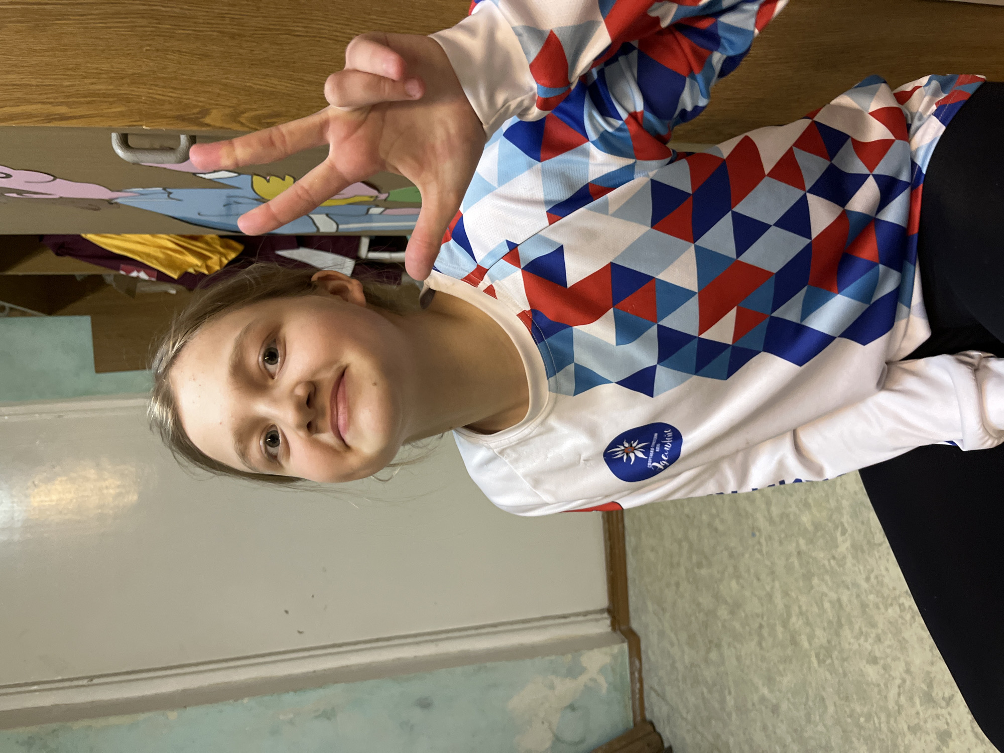 A young girl is posing indoors, making a hand gesture. She is wearing a long-sleeved shirt with a geometric pattern of red, white, and blue triangles and a blue patch with an emblem on the chest. Her hand is raised near the top of the frame, making a symbol. The background includes a painted wooden cabinet and wall. The photo may be taken in an interior in Berezniki, Russia, as the emblem on her shirt is similar to the emblem of a sports organization in that city.