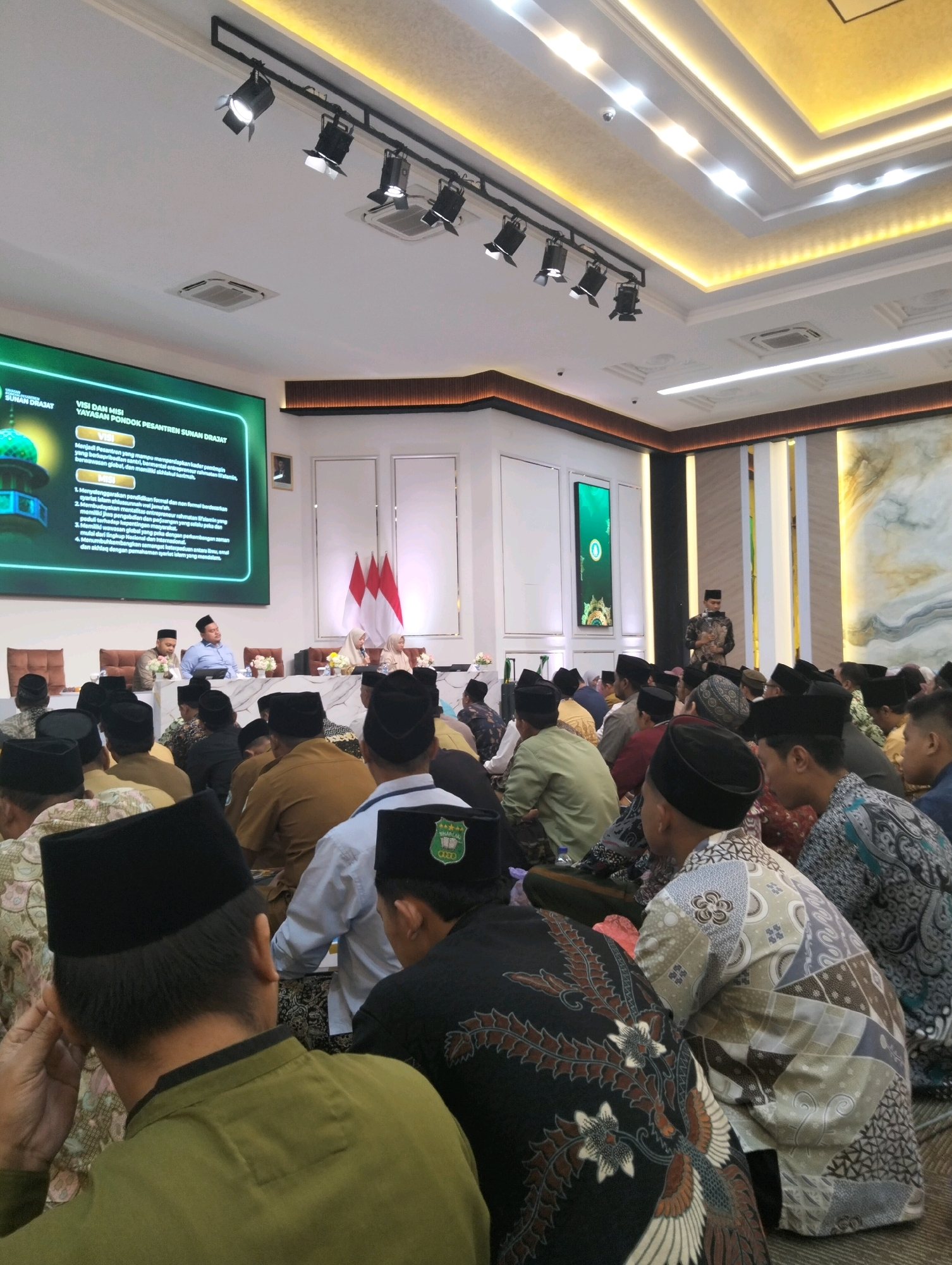 The image depicts an indoor assembly, likely a formal event or meeting, taking place in a well-lit room with a modern ceiling featuring recessed lighting and spotlights. In the foreground, a crowd of men, mostly wearing traditional Indonesian attire including peci (caps), are seated on the floor facing a stage area. The men are diverse in age and attire, with some wearing batik shirts and others plain colored shirts.
The stage area features a large screen displaying text in Indonesian, with the title "YAYASAN PONDOK PESANTREN SUNAN DRAJAT" (Sunan Drajat Islamic Boarding School Foundation) and sections for "VISI DAN MISI" (Vision and Mission). Three Indonesian flags are displayed on the stage. Several individuals, including men and women, are seated at a table on the stage, appearing to be speakers or honored guests. One man in the center of the stage is speaking.
The overall environment suggests a place of learning or religious gathering, possibly within the Sunan Drajat Islamic Boarding School campus in Paciran, Lamongan Regency, East Java, Indonesia. The presence of the foundation's name and the nature of the gathering strongly point to this location. The room is spacious, with additional decorative elements like vertical wooden panels and a large abstract artwork on a far wall. The atmosphere appears attentive and formal.