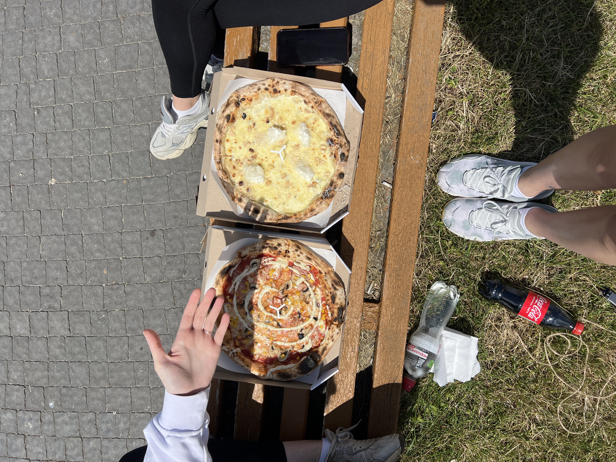 In Ternopil, Ukraine, two pizzas rest in their cardboard boxes on a wooden bench. One pizza is topped with red sauce, cheese, mushrooms, corn, and a white creamy swirl, while the other features a white sauce base, cheese, and dollops of a soft white cheese. Beside the pizzas, a black smartphone lies on the bench. On the grass below the bench, a partially consumed bottle of Coca-Cola and a clear bottle of water are visible, along with a stack of white napkins and a spooled piece of twine. The scene is set against a backdrop of gray paving stones and green grass, suggesting an outdoor public space. Two individuals are partially visible: one's legs and white sneakers are in the foreground, and another's hand, wearing a ring, extends over the pizza box on the left, indicating shared consumption of the food. The lighting suggests a sunny day.