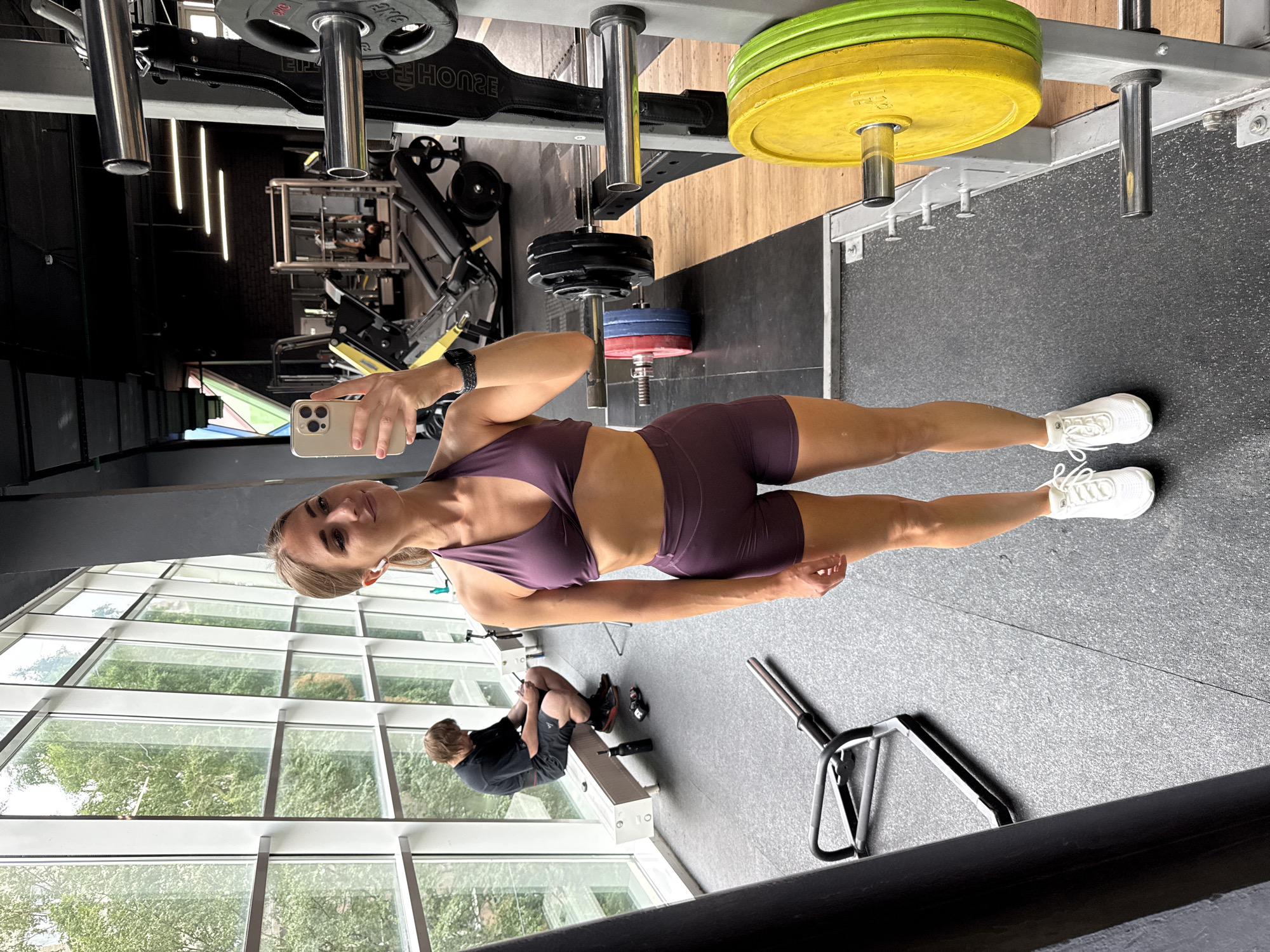 The image depicts a young woman taking a selfie in a gym. She is wearing a purple sports bra and matching shorts, along with white athletic shoes and wireless earbuds. She is holding a smartphone in her right hand. In the background, another person is sitting on a bench, looking at their phone. The gym appears to be well-lit with large windows overlooking a leafy area, suggesting it might be located in a park or a green district of Saint Petersburg, Russia, possibly within a modern building with ample natural light. The gym equipment visible includes weight plates, barbells, and a weight machine. The overall atmosphere is one of fitness and activity.