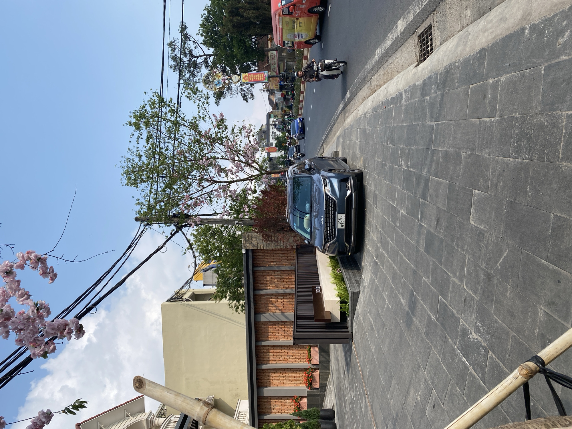The image depicts a street scene in Da Lat, Vietnam. A dark blue SUV is parked on a tiled sidewalk next to a building with a brick facade and a black metal frame. The license plate of the SUV is visible. The building's architectural details include planted boxes with greenery and red flowers. In the background, a street is visible with various vehicles including a red van, motorcycles, and cars moving along. Trees with light pink blossoms line the street. Overhead, power lines are visible. The sky is blue with some clouds. There's a decorative floral display mounted on a pole in the background.